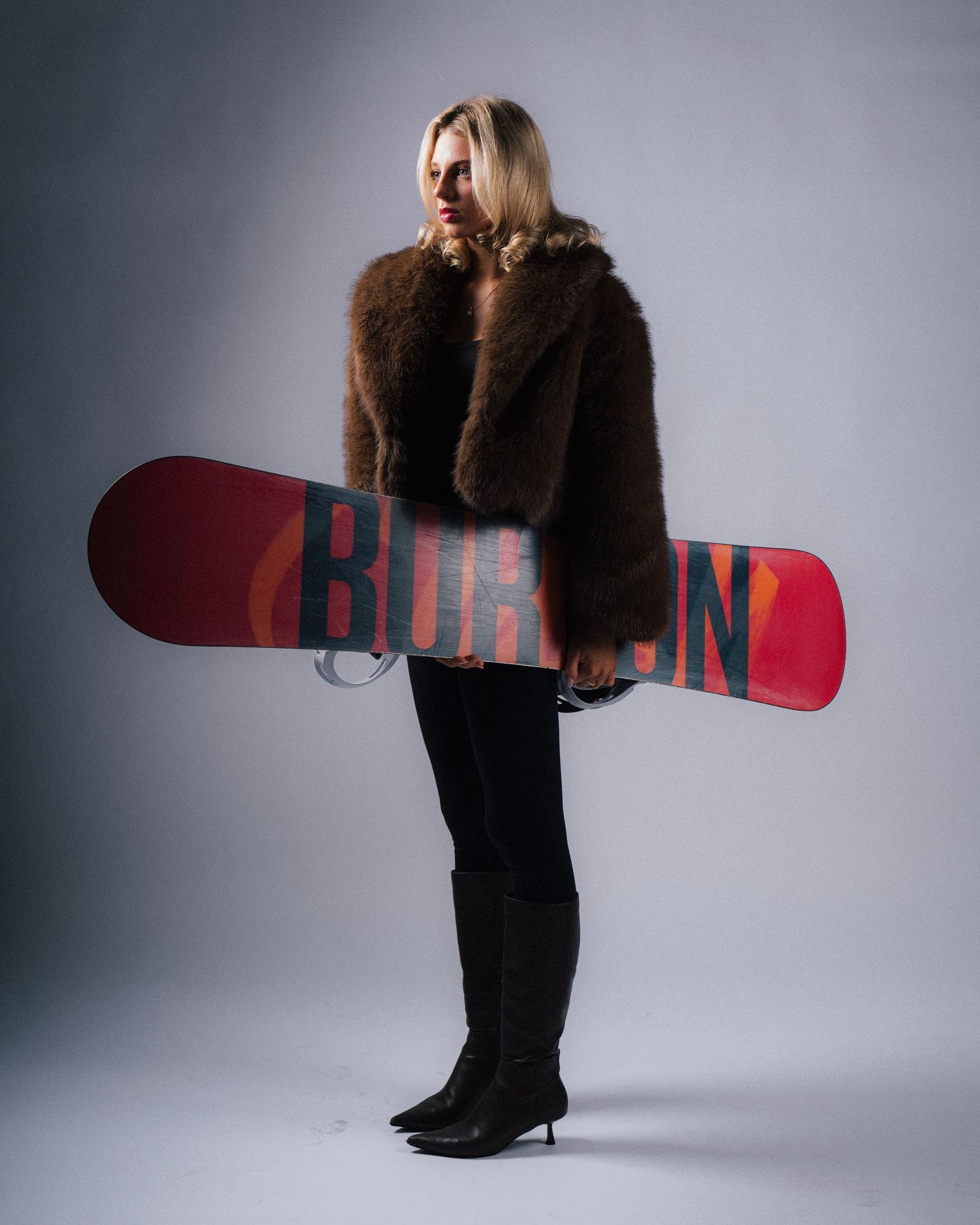 Snowboarder in a brown fur coat and black boots holds a red Burton snowboard against a gray backdrop.