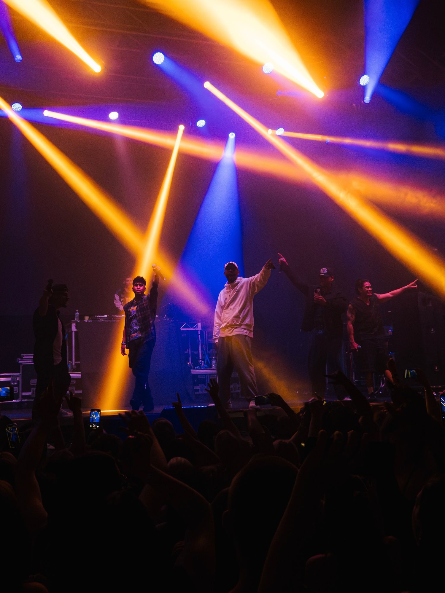 Performers silhouetted on stage with dramatic orange and blue spotlights creating dynamic beams during a concert performance.