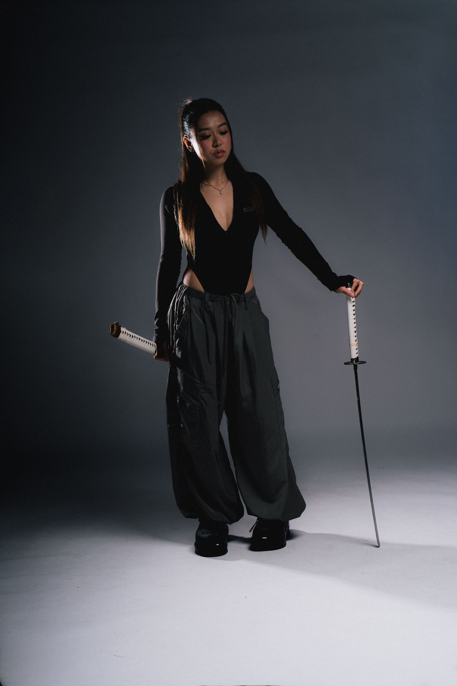 Dramatic portrait of person in black outfit holding sword against grey background.
