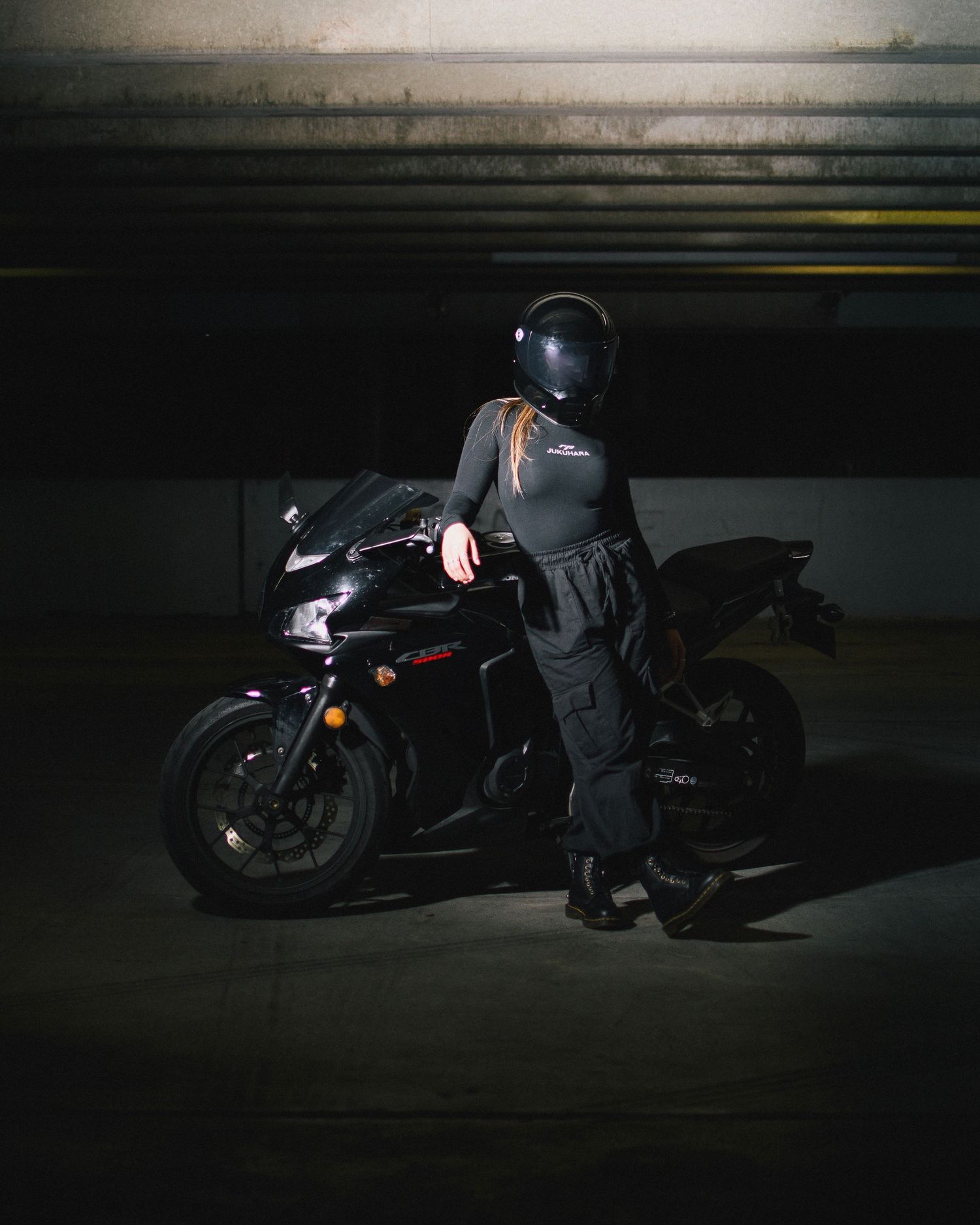Motorcyclist in black protective gear standing beside sport bike in dark parking garage.