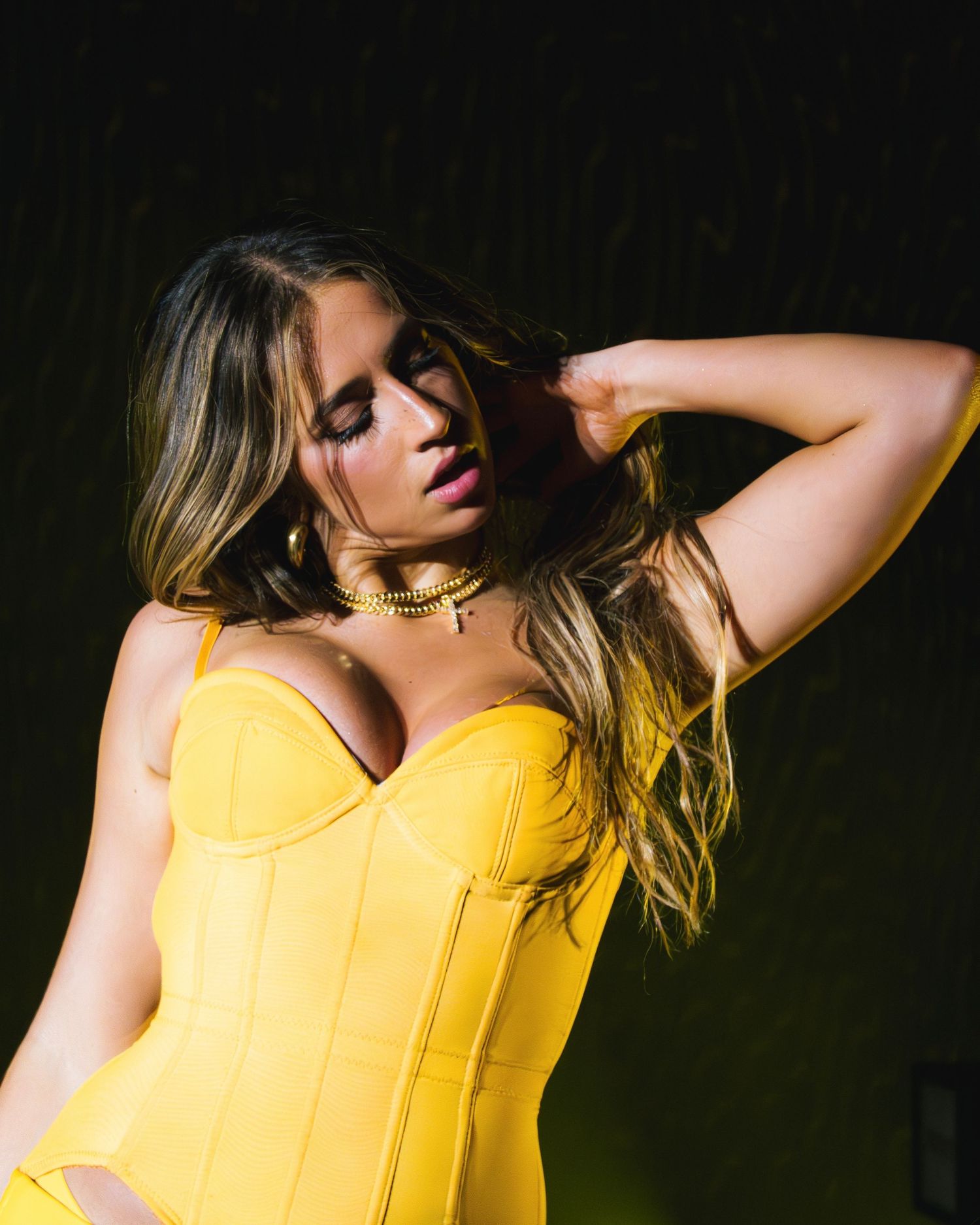 Model in yellow corset poses elegantly against dark background with dramatic lighting.