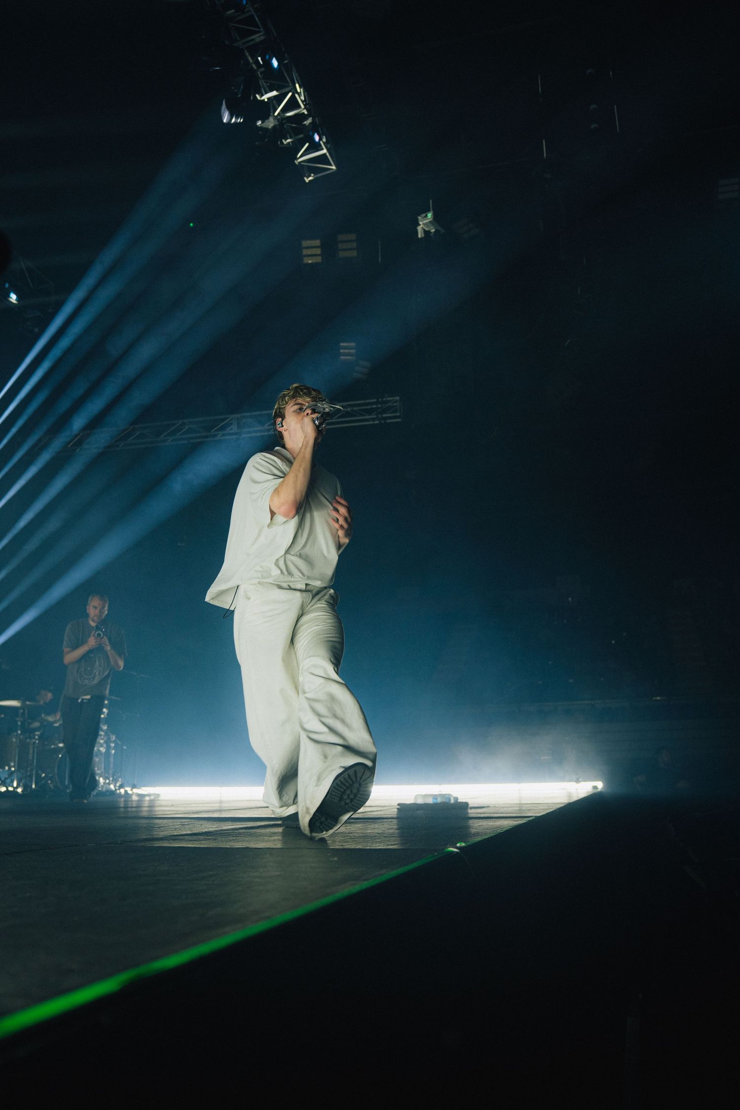 Performer in white outfit stands on stage with dramatic blue spotlights and beams illuminating the dark concert venue.