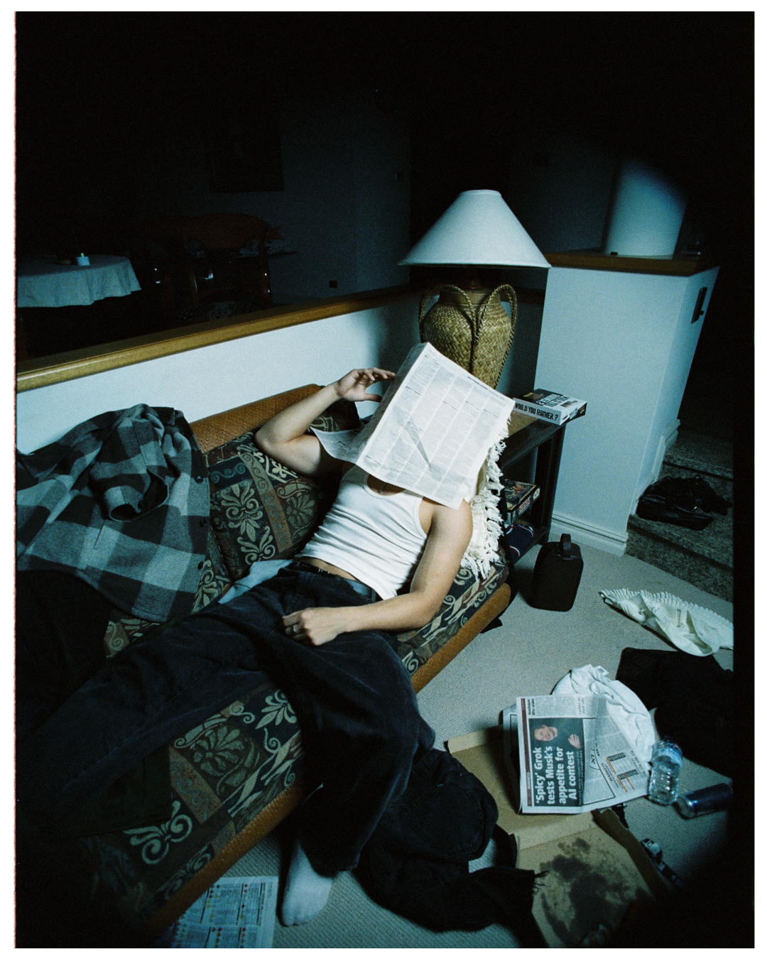 Someone lies relaxed on a patterned couch reading a book in a dimly lit room with a table lamp casting blue light.