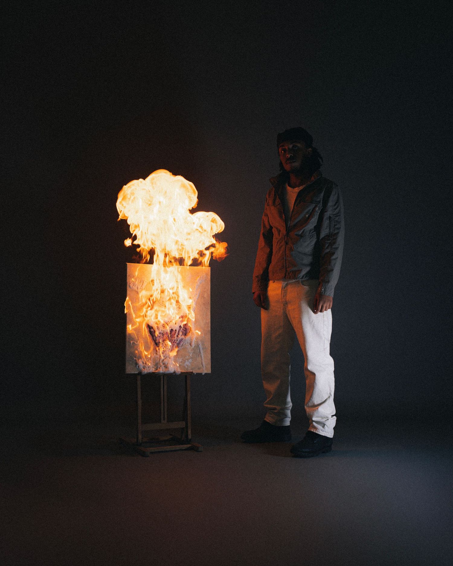A dramatic dark scene with a flaming fire box sculpture on a stand next to someone in casual clothing.