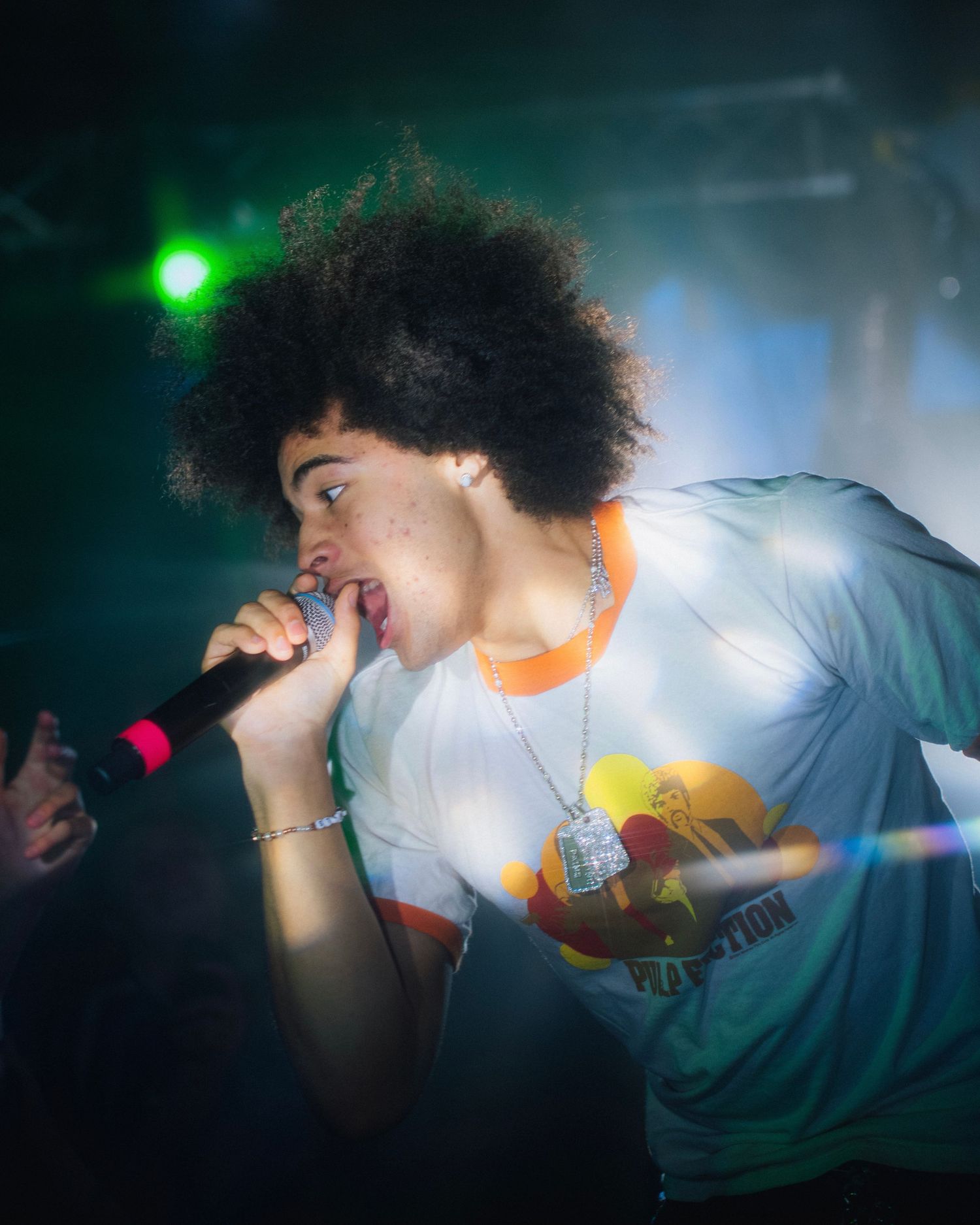 Energetic performer with afro hairstyle singing into microphone during concert with colorful lighting.