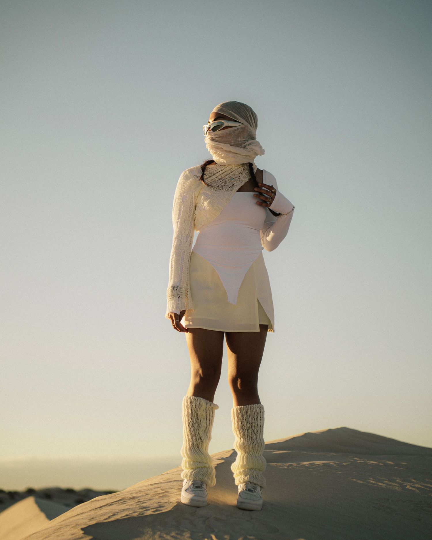 Figure in a white dress and leg warmers standing on a sand dune against a bright sky.