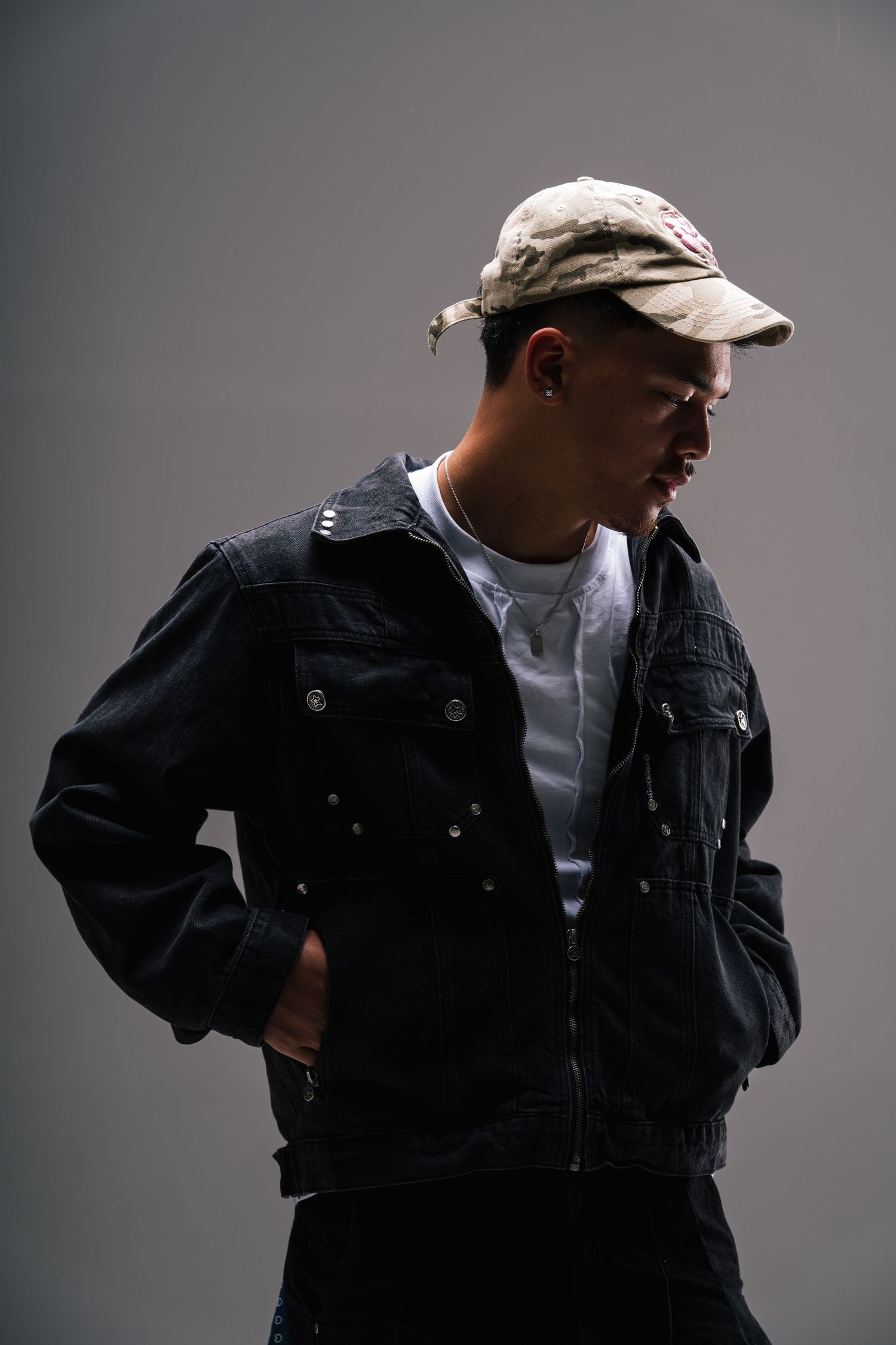 Side profile of person in dark denim jacket and baseball cap against gray background in dramatic lighting.