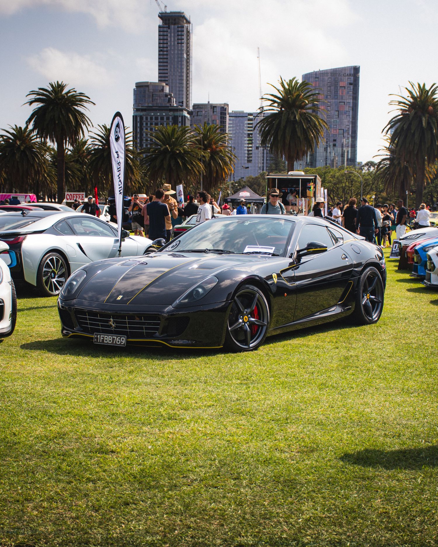 Black Ferrari sports car on display at outdoor car show with palm trees and skyscrapers.