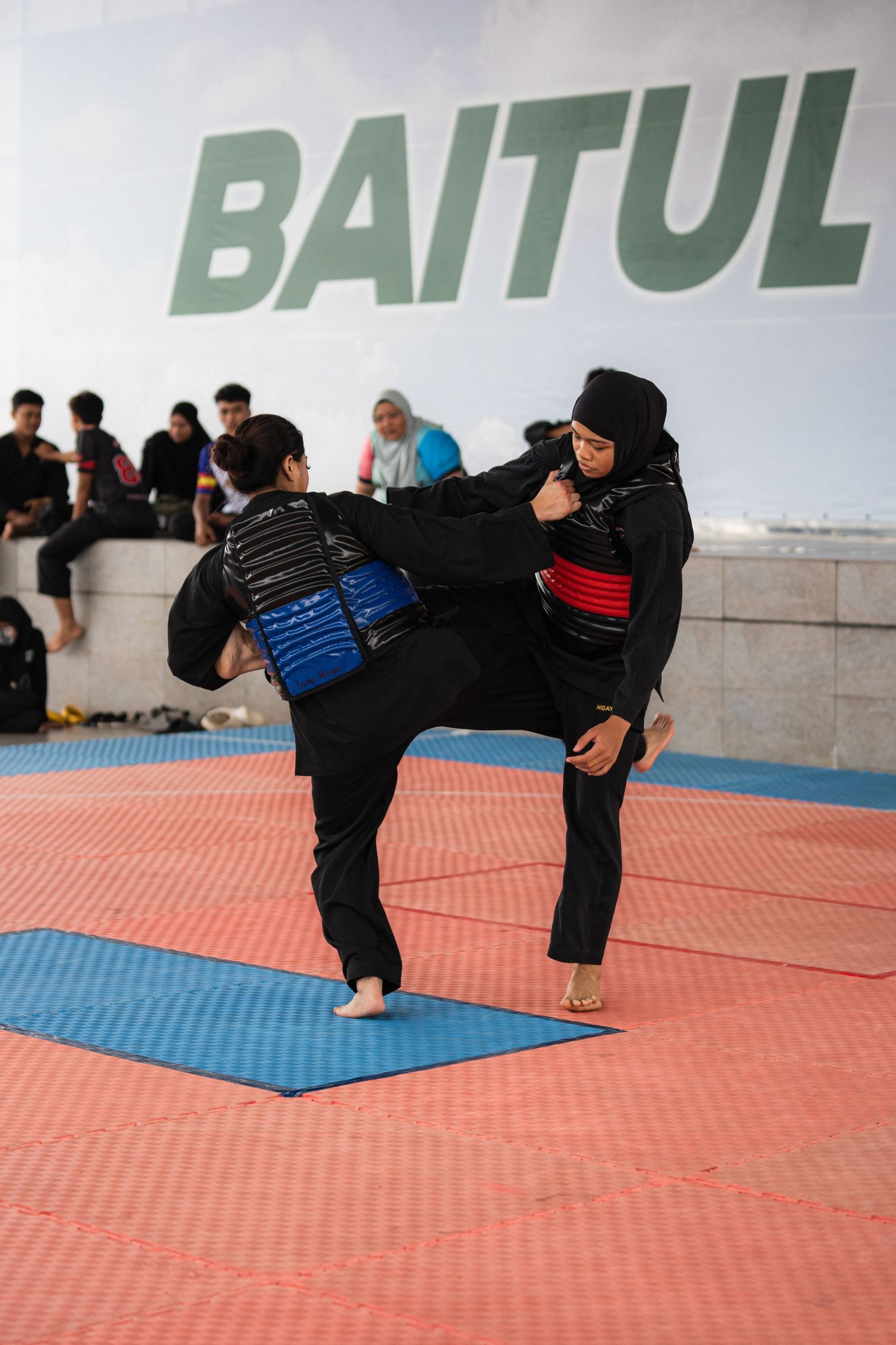 Martial arts practitioners demonstrate kicking techniques under a Baitul sign on red mats.