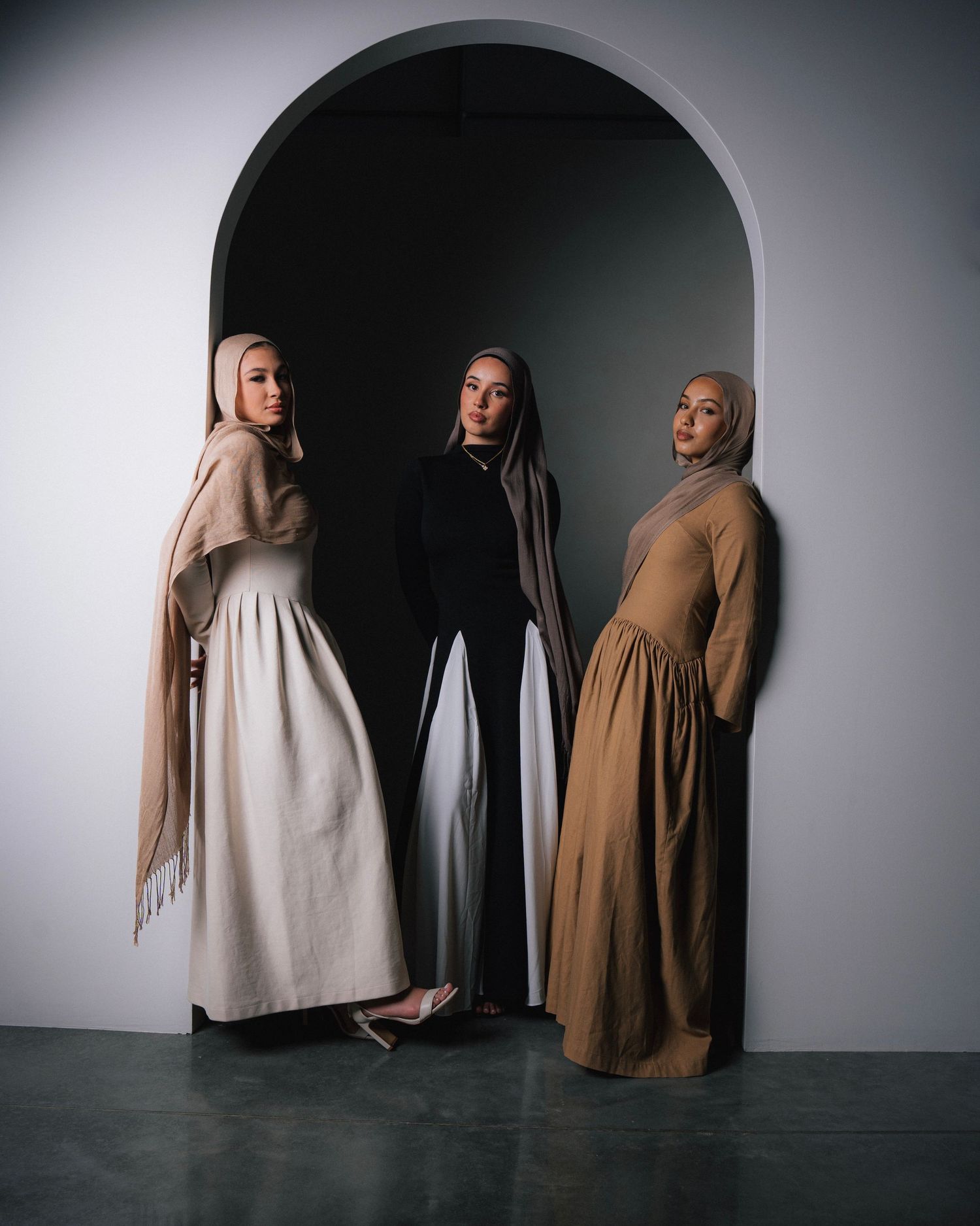 Three women in flowing maxi dresses pose elegantly in front of arched doorway.