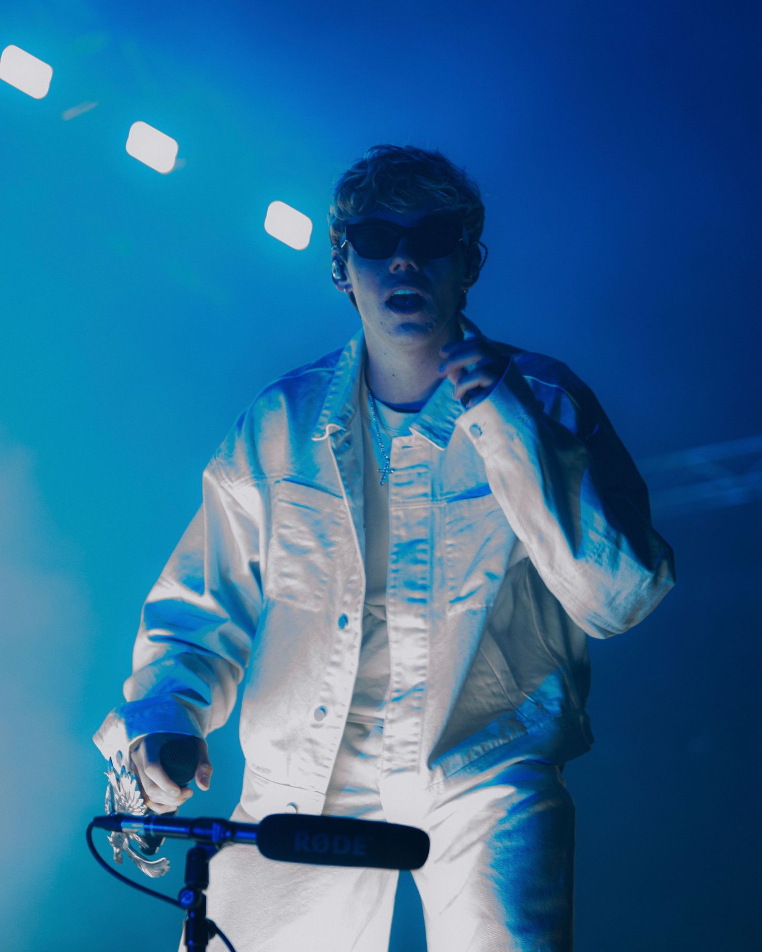 Concert performer in white denim jacket illuminated by blue stage lights while holding DJ equipment.