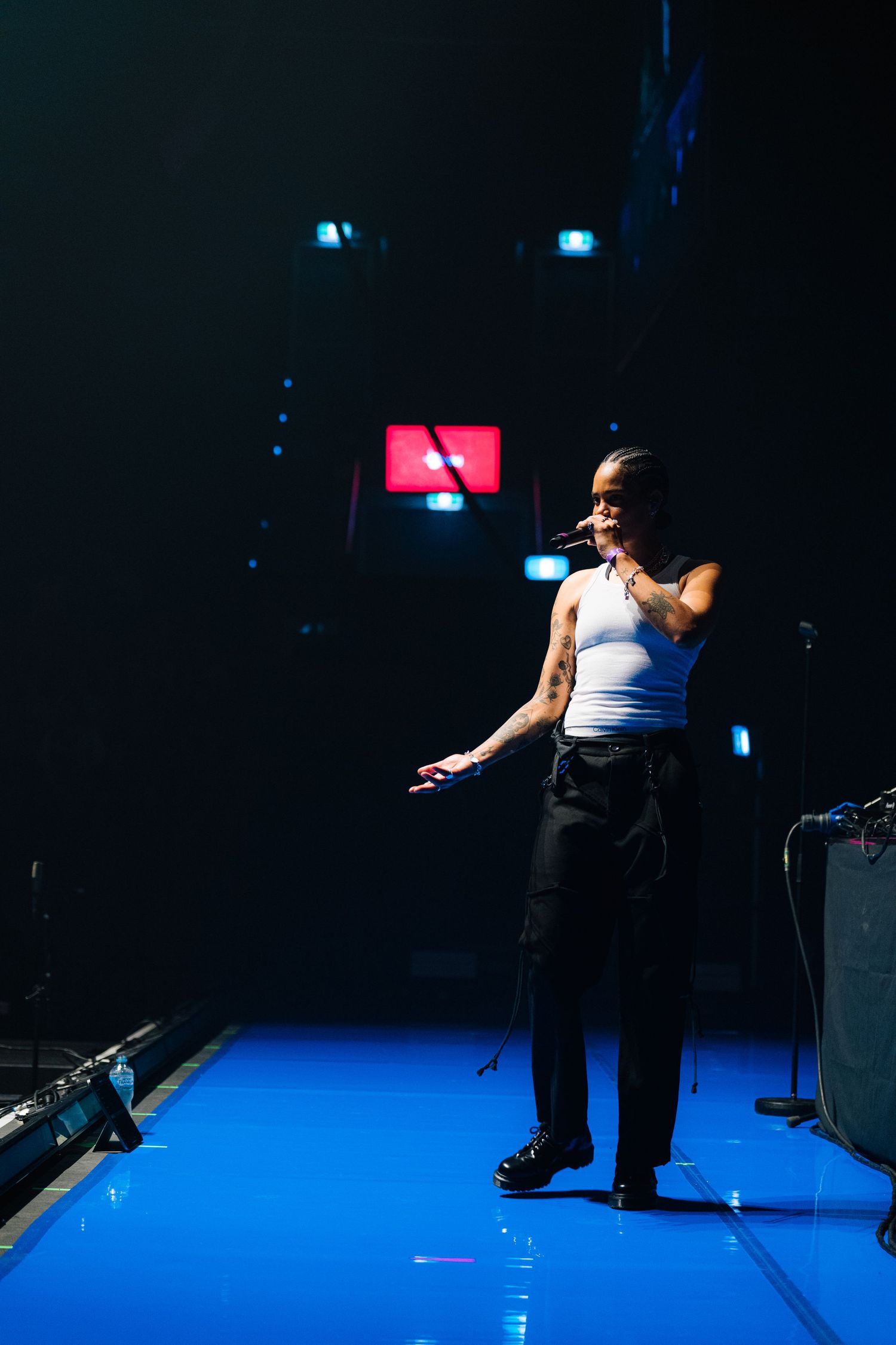 Performer in white tank top and dark pants stands on illuminated blue stage with dramatic lighting and sound equipment.