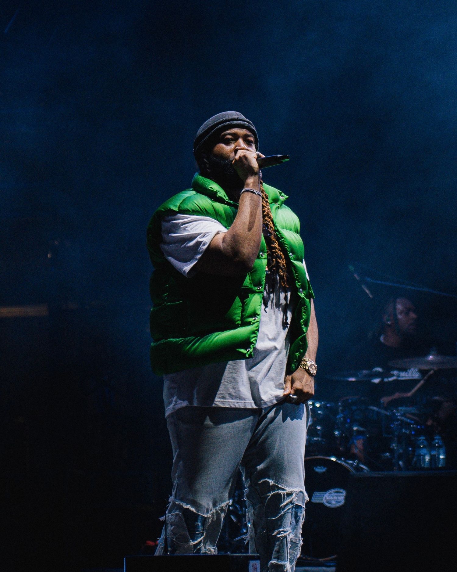 Performer in green and white vest outfit singing into microphone on dimly lit stage.