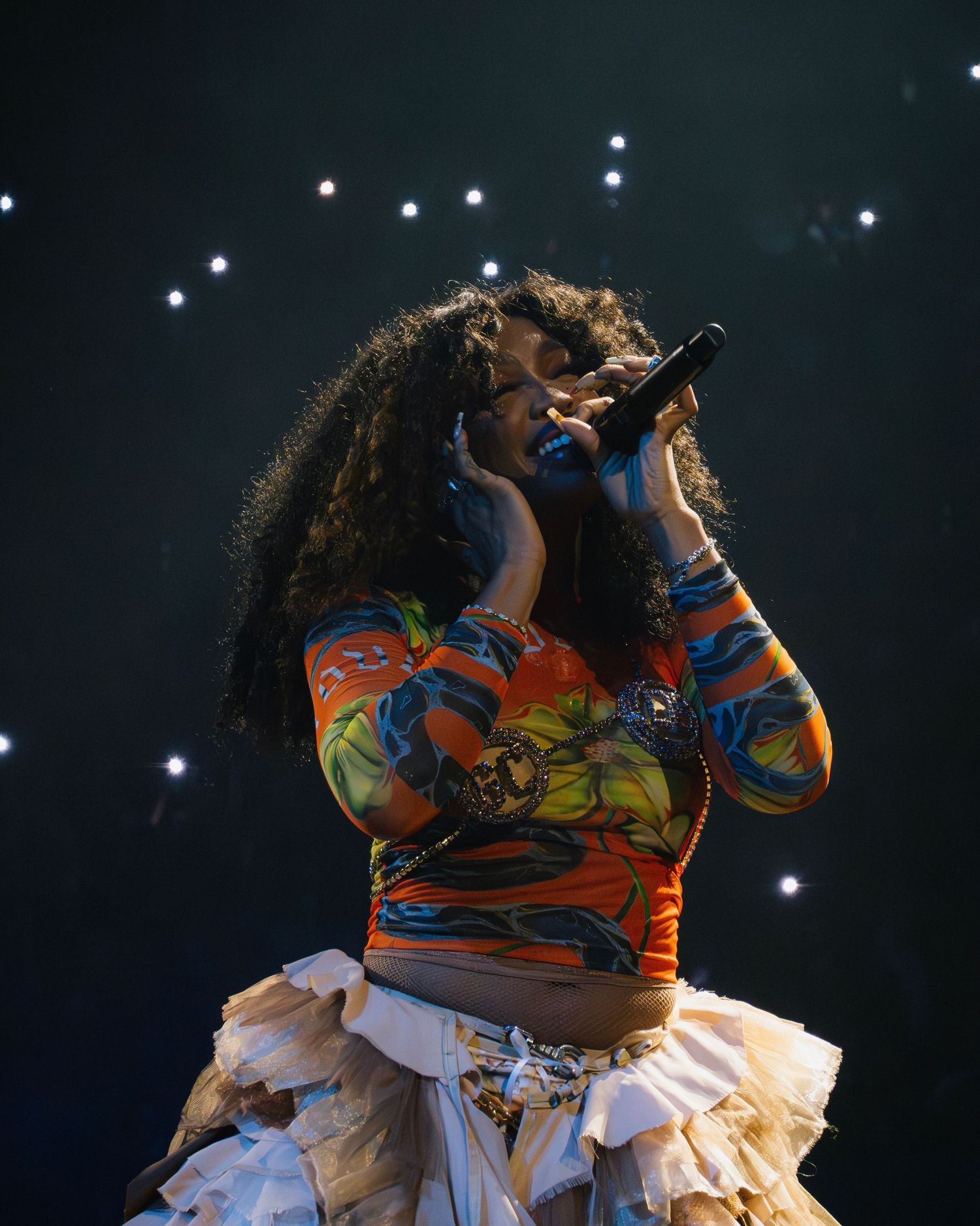 Performer in a colorful outfit singing on stage with spotlights creating a dramatic backdrop.