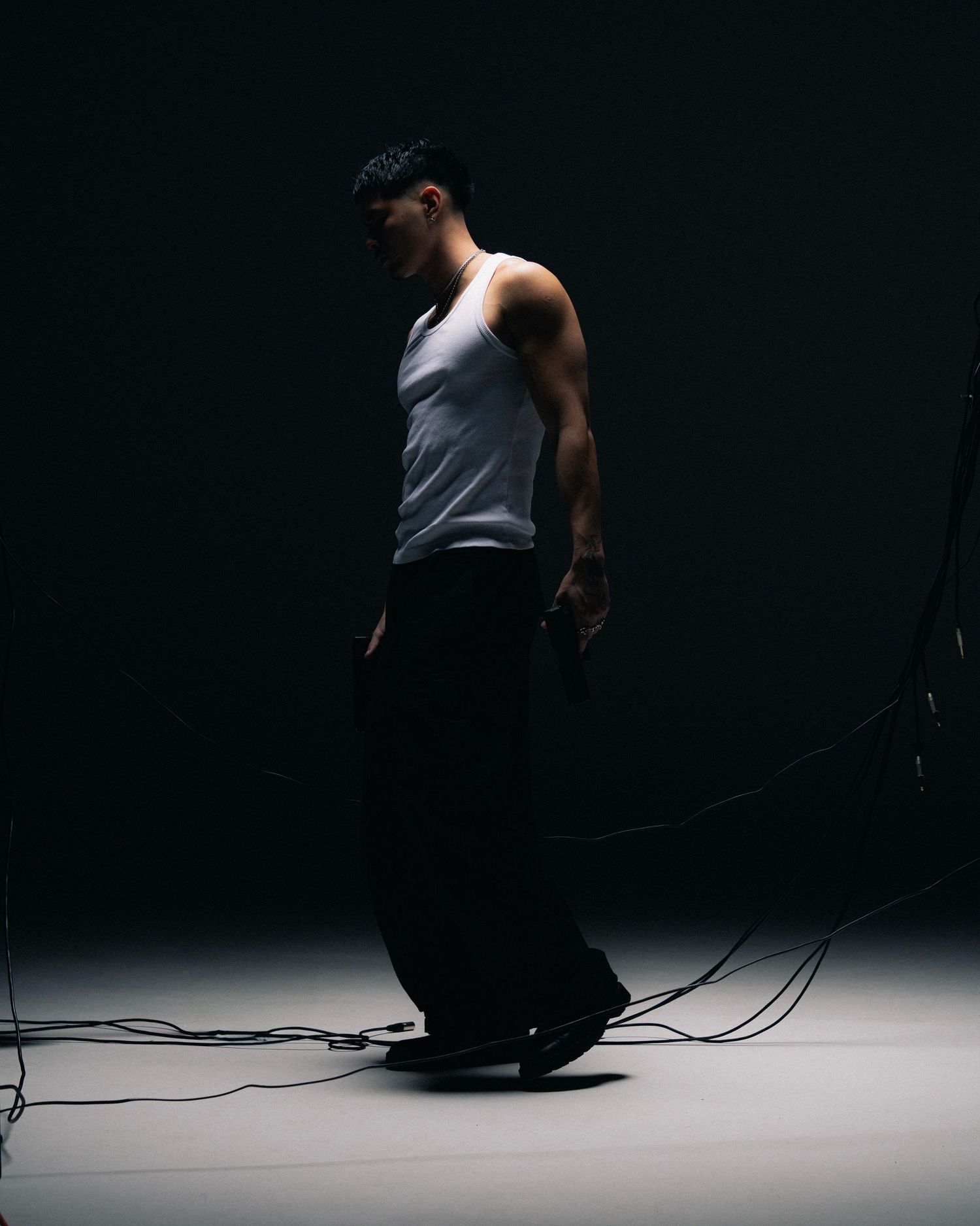 Figure in white tank top and dark pants stands dramatically in low light against dark background with cables on floor.