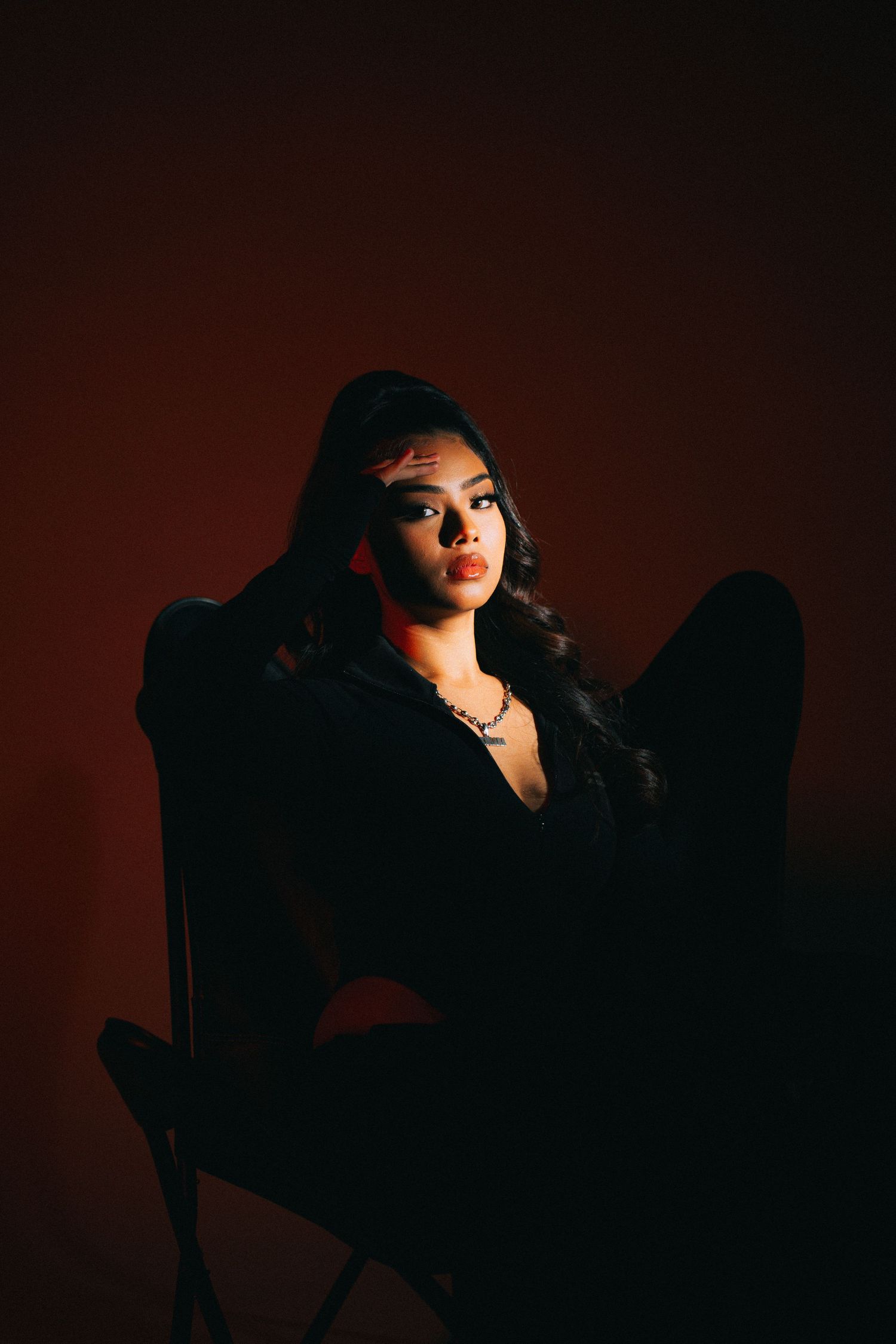 Moody portrait of someone seated in a dark chair against a red backdrop with dramatic lighting and shadows.