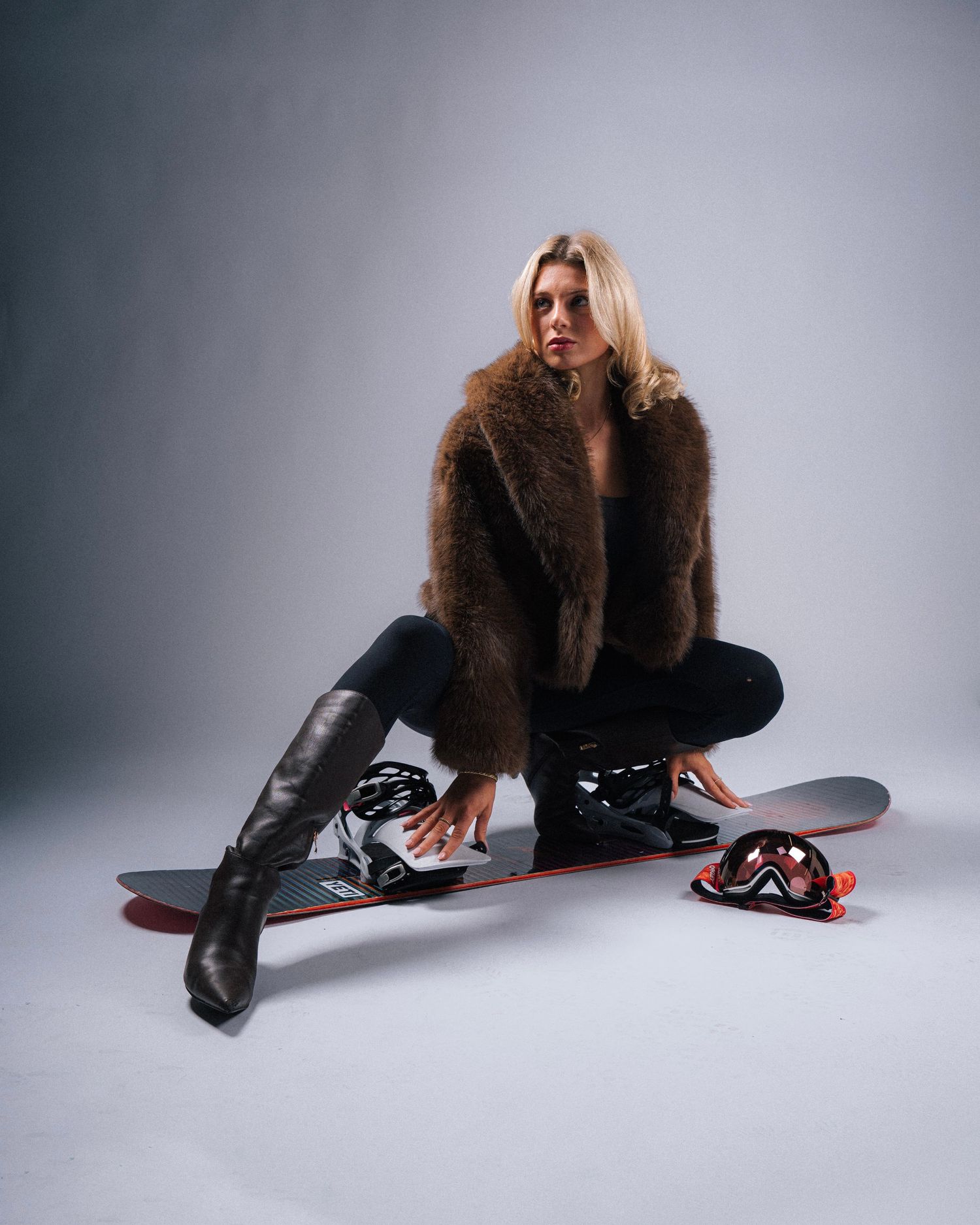 Fashion model sits on a snowboard wearing a brown fur coat and tall black boots in a studio setting.