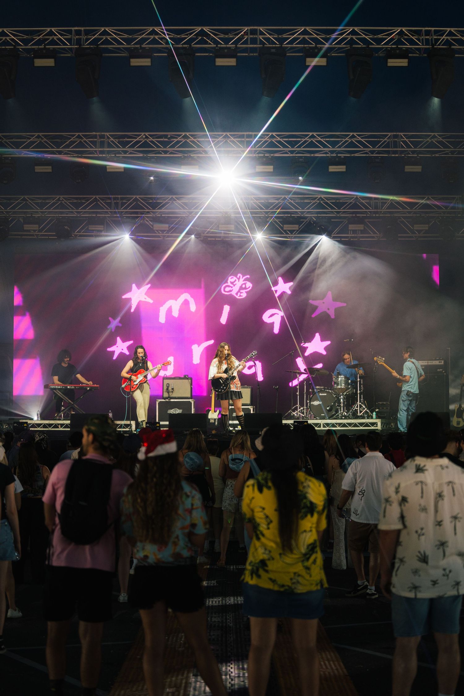 Concert crowd watches performers on stage with pink lighting and bright spotlights creating an energetic atmosphere.