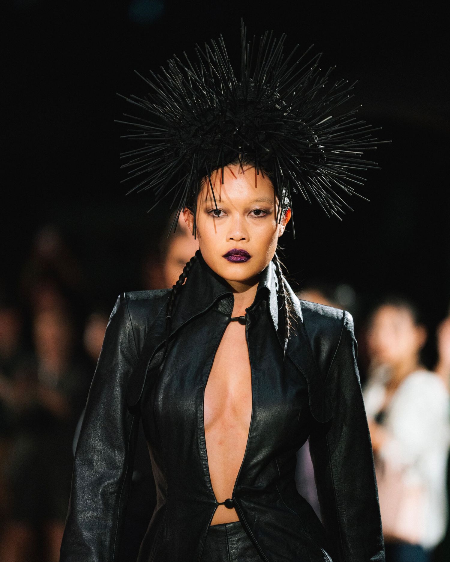 Fashion model wearing dramatic spiked headpiece and black leather outfit on runway.