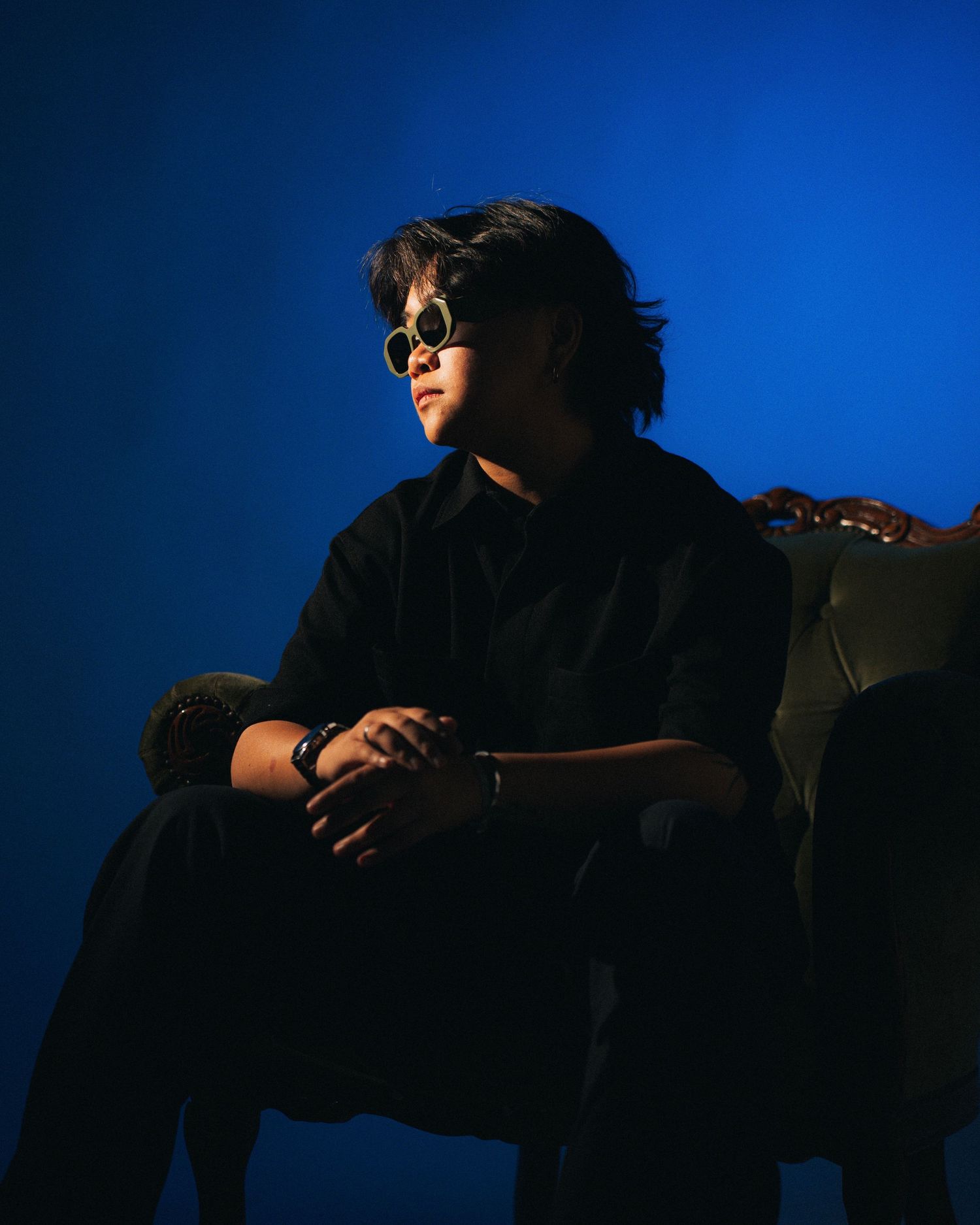 Silhouetted figure wearing sunglasses sits on couch against vibrant blue backdrop.
