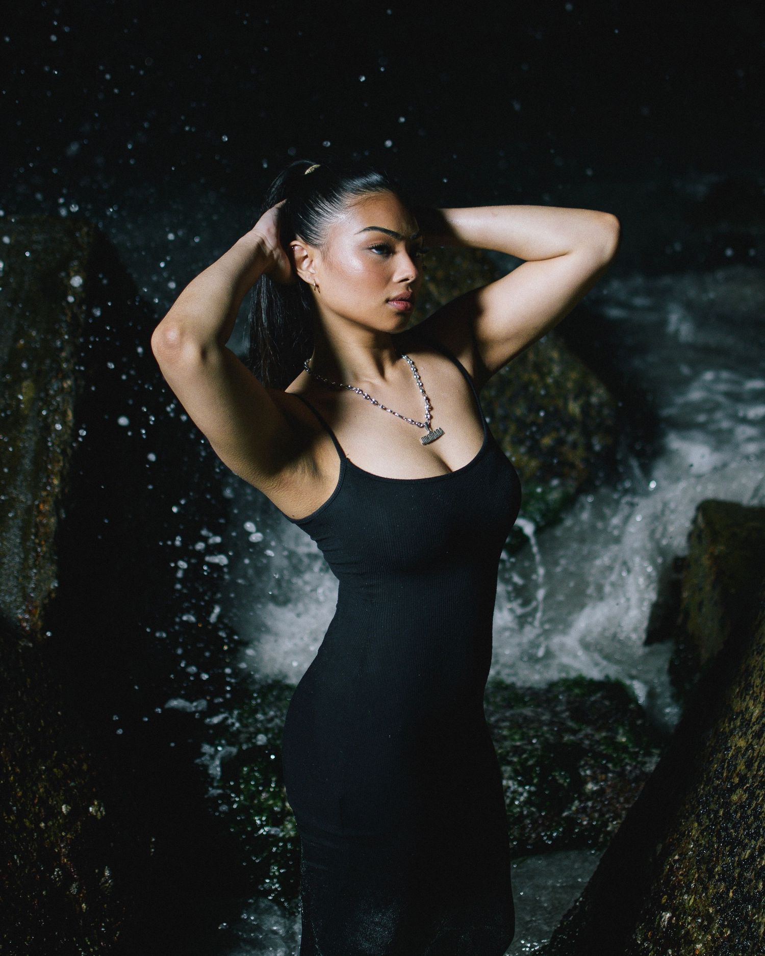 A dramatic fashion portrait in a black dress against a dark waterfall backdrop with moody lighting.