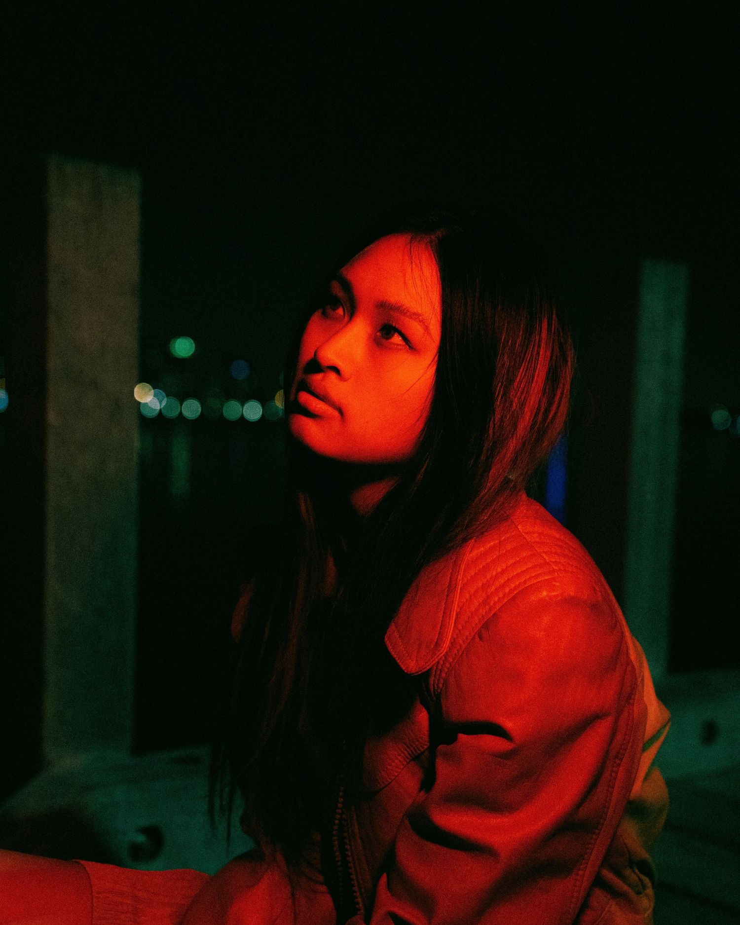 A figure captured in moody red lighting at night appears pensive while wearing a casual jacket against a dark background.