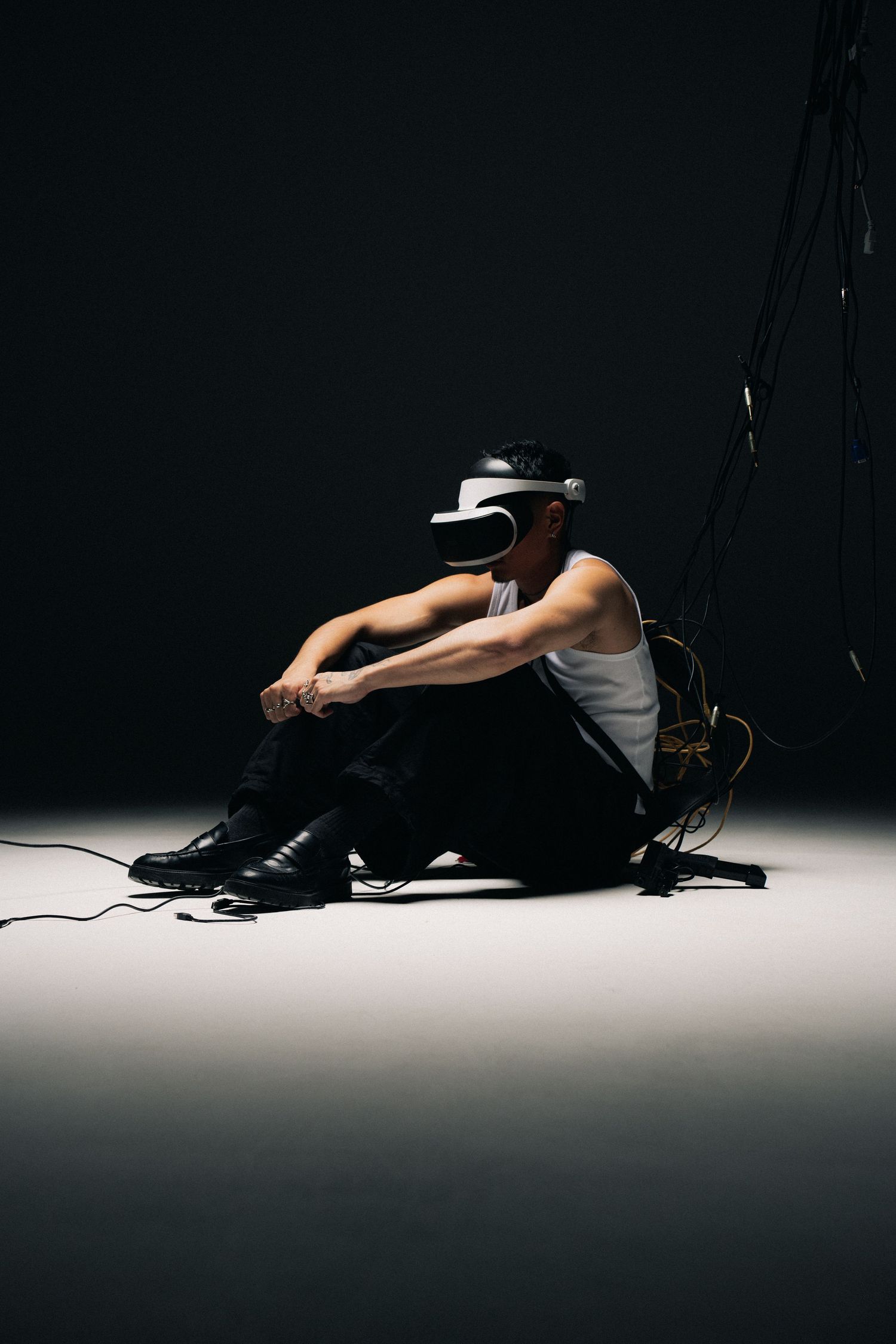 Figure in white tank top and black pants sits on floor against dark background wearing VR headset with cables.