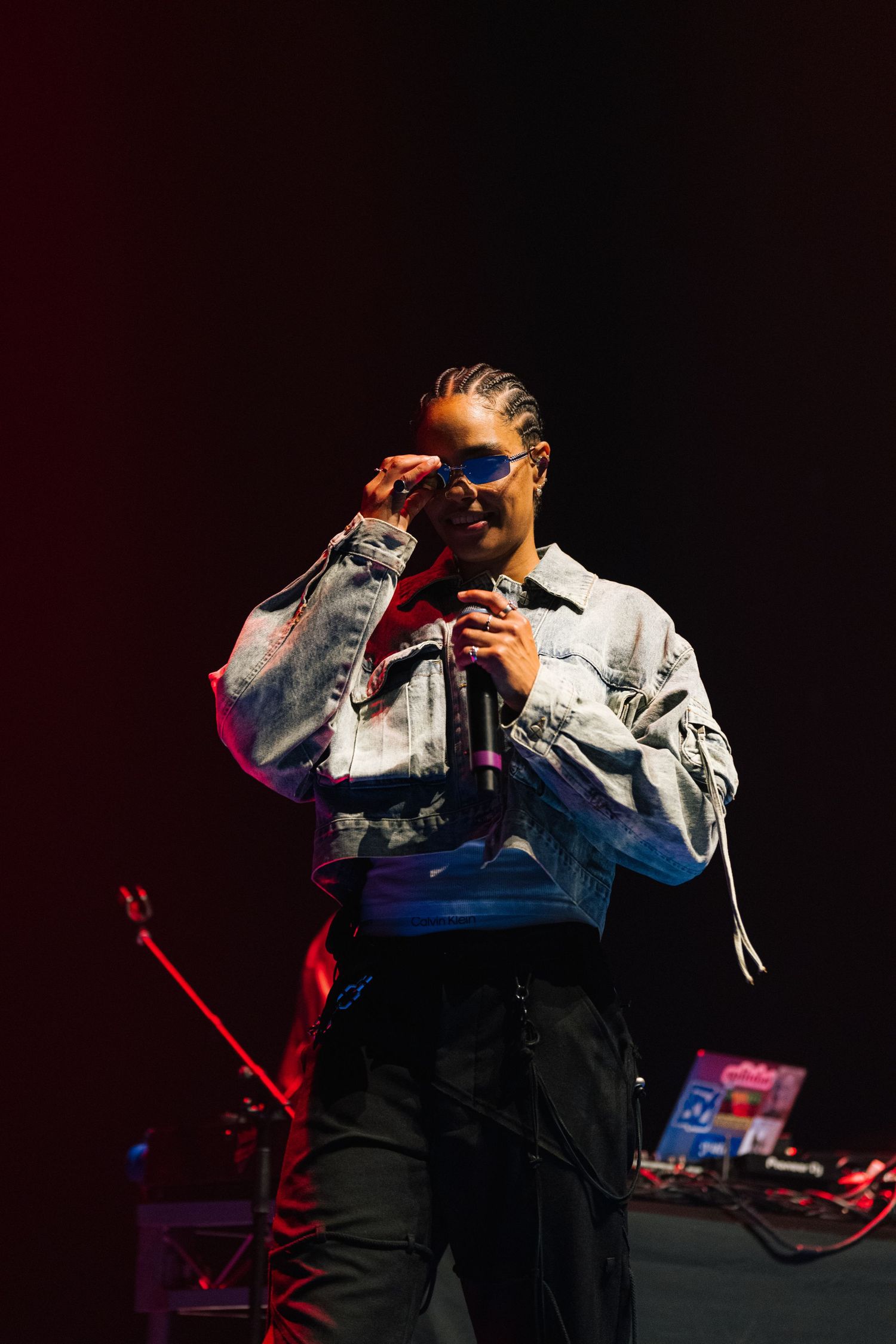 A performer in denim jacket and dark pants sings on stage with dramatic lighting and music equipment in the background.