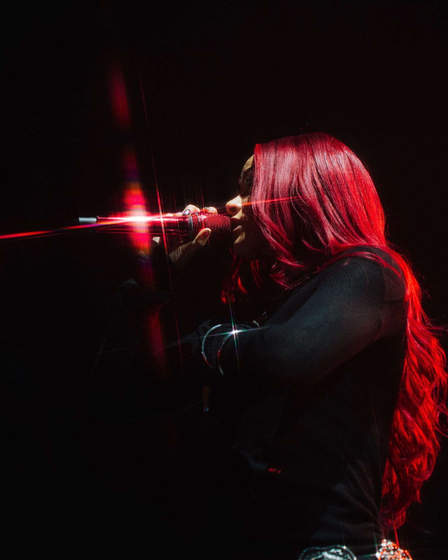 A silhouetted performer with long red hair sings into a microphone on a dark stage with dramatic red lighting.