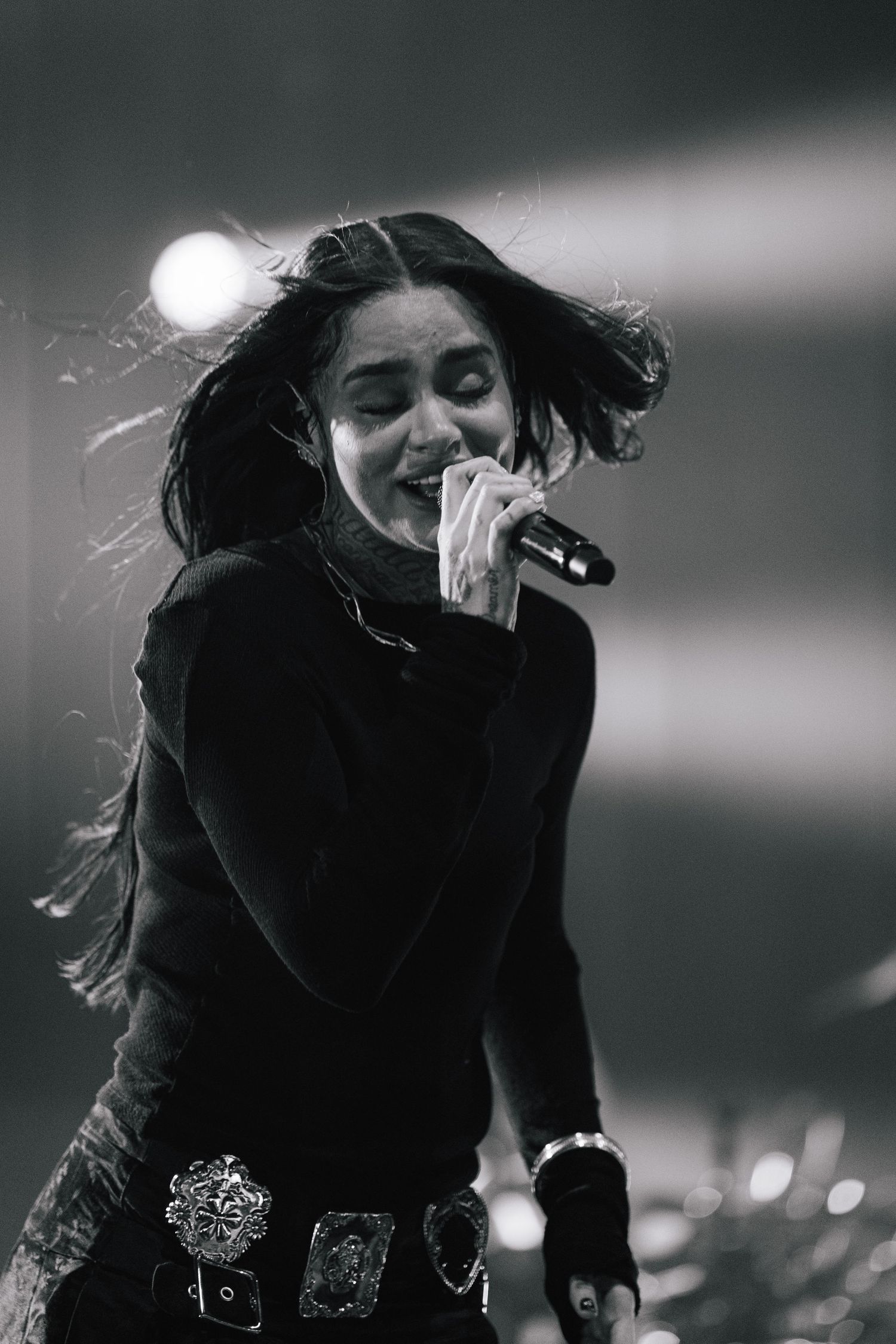 Black and white concert photo of performer singing passionately in black outfit with flowing hair and statement belt.