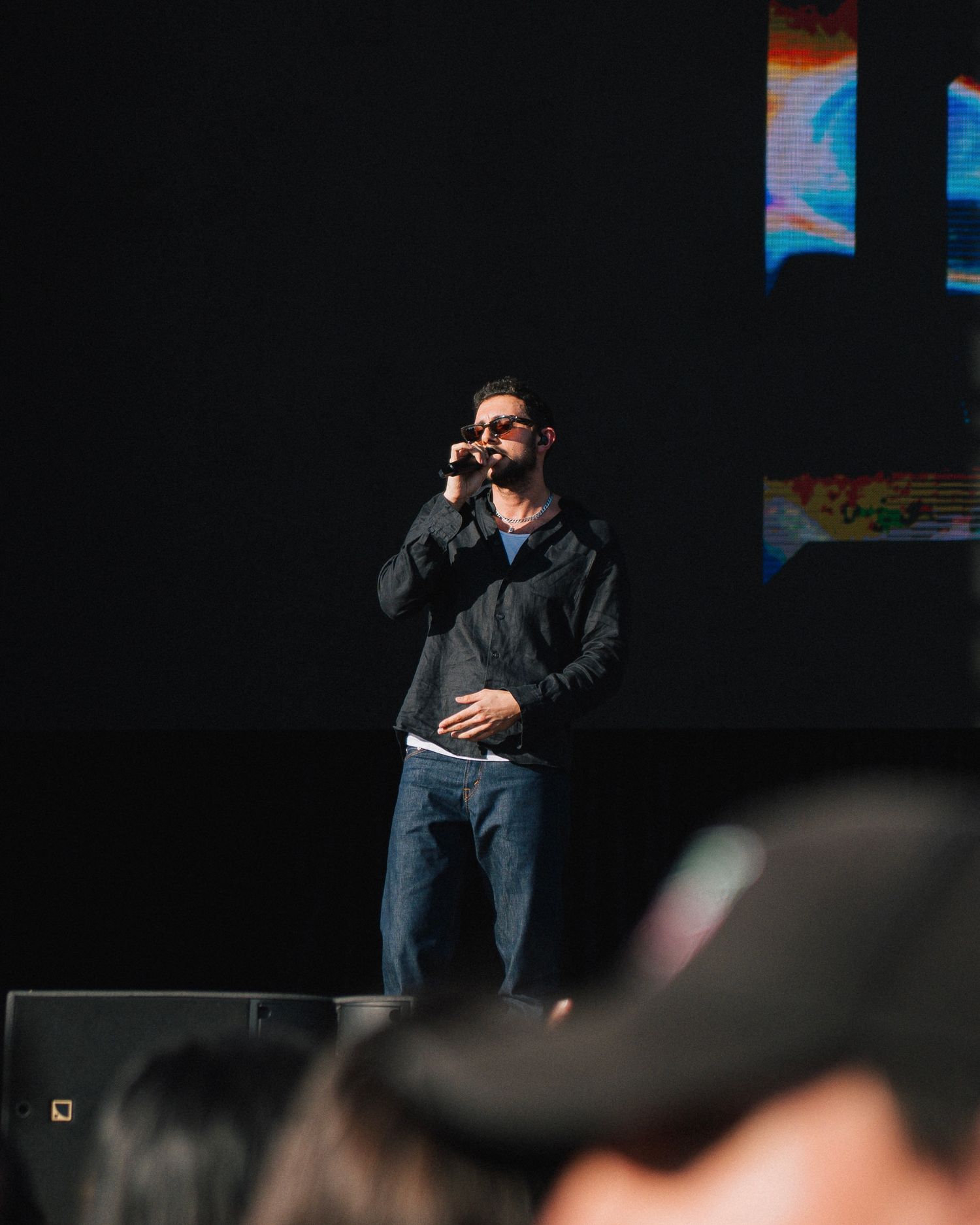 Performer in casual attire speaks into microphone on dimly lit stage.