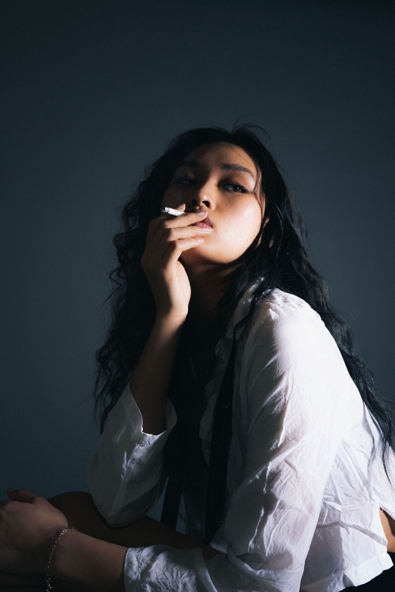 Figure in a flowing white blouse strikes a dramatic pose against dark background while smoking a cigarette.