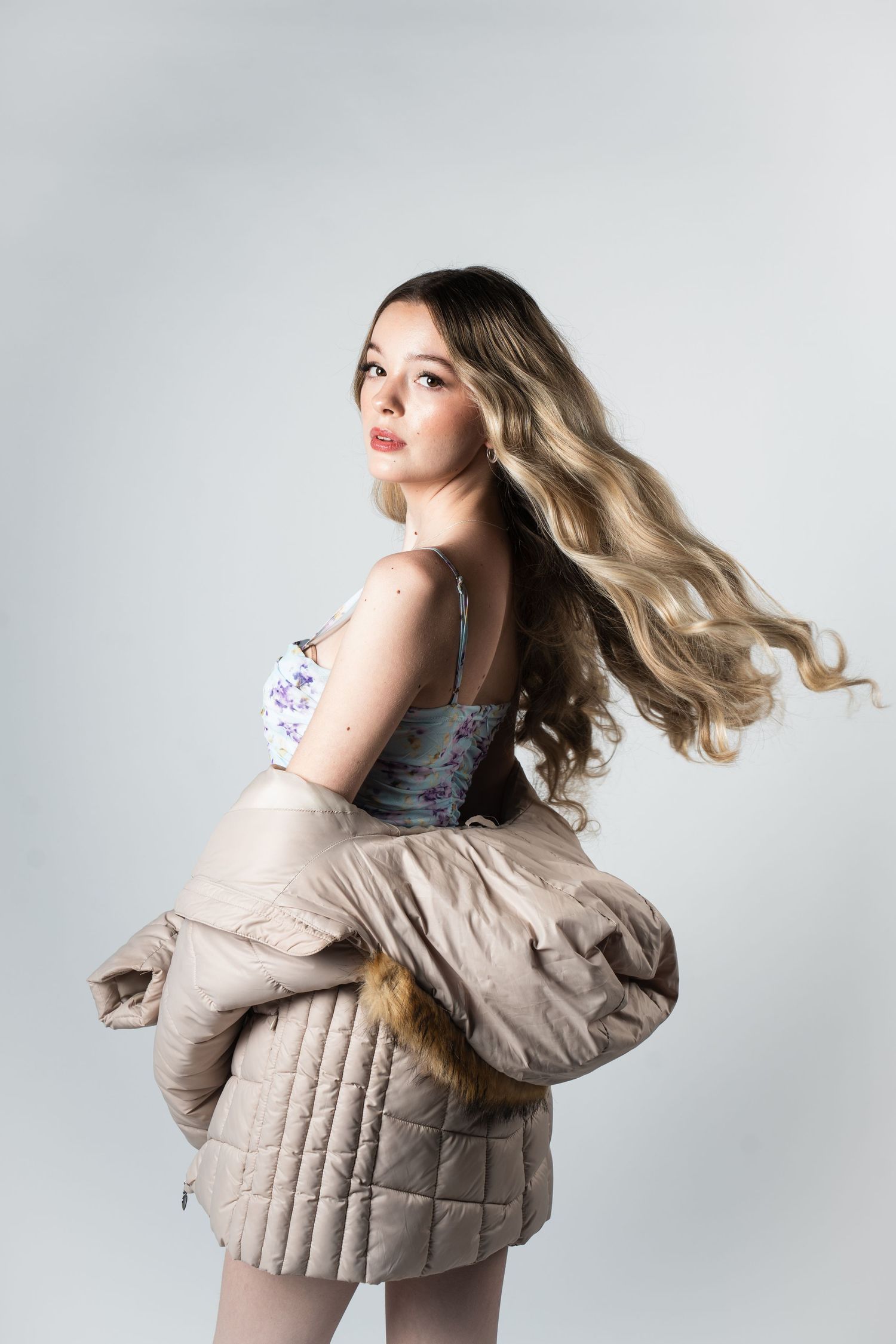 Model in floral dress with flowing blonde hair twirls while holding beige quilted puffer jacket against white background.