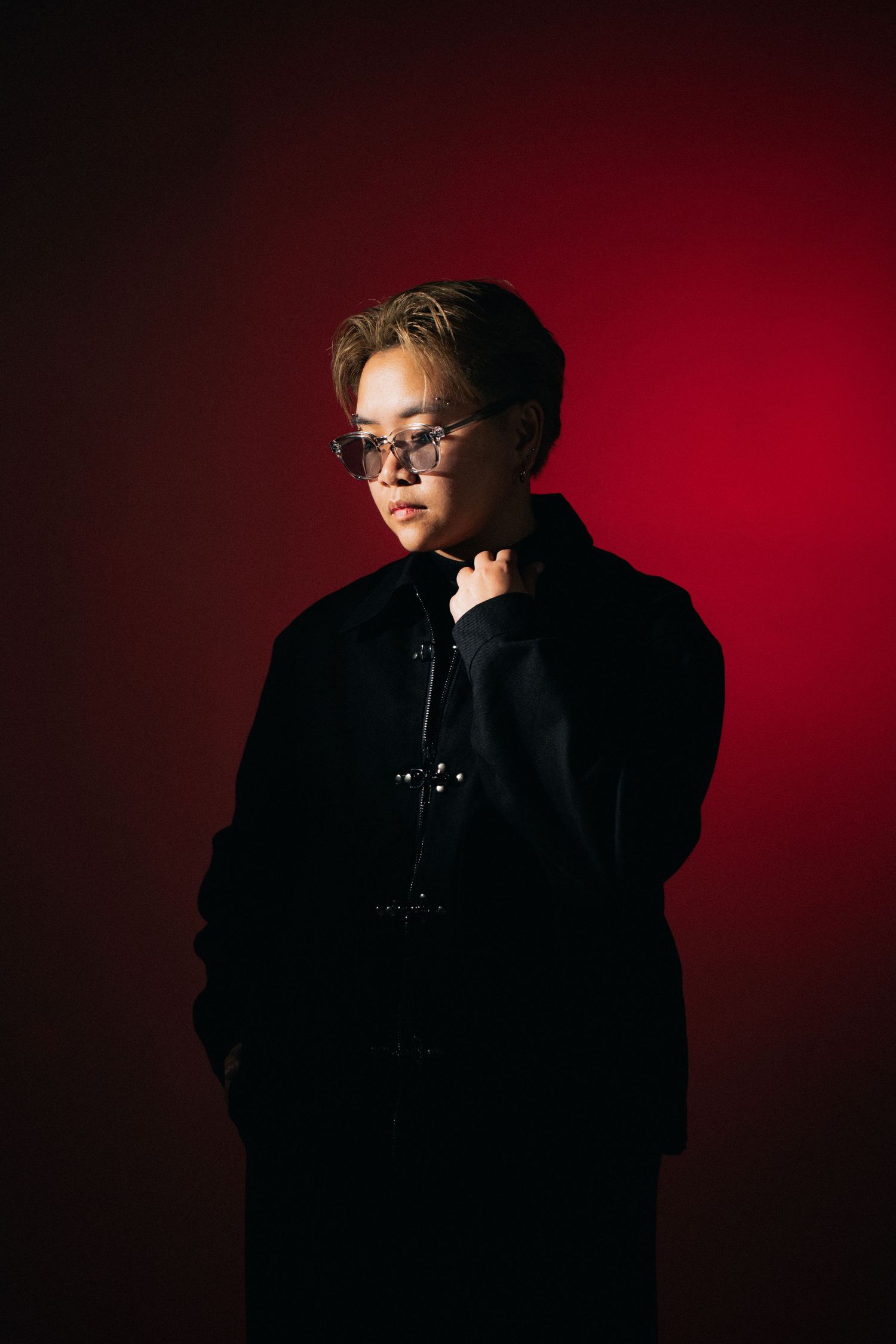 A figure in black winter coat and glasses poses dramatically against a deep red studio backdrop.