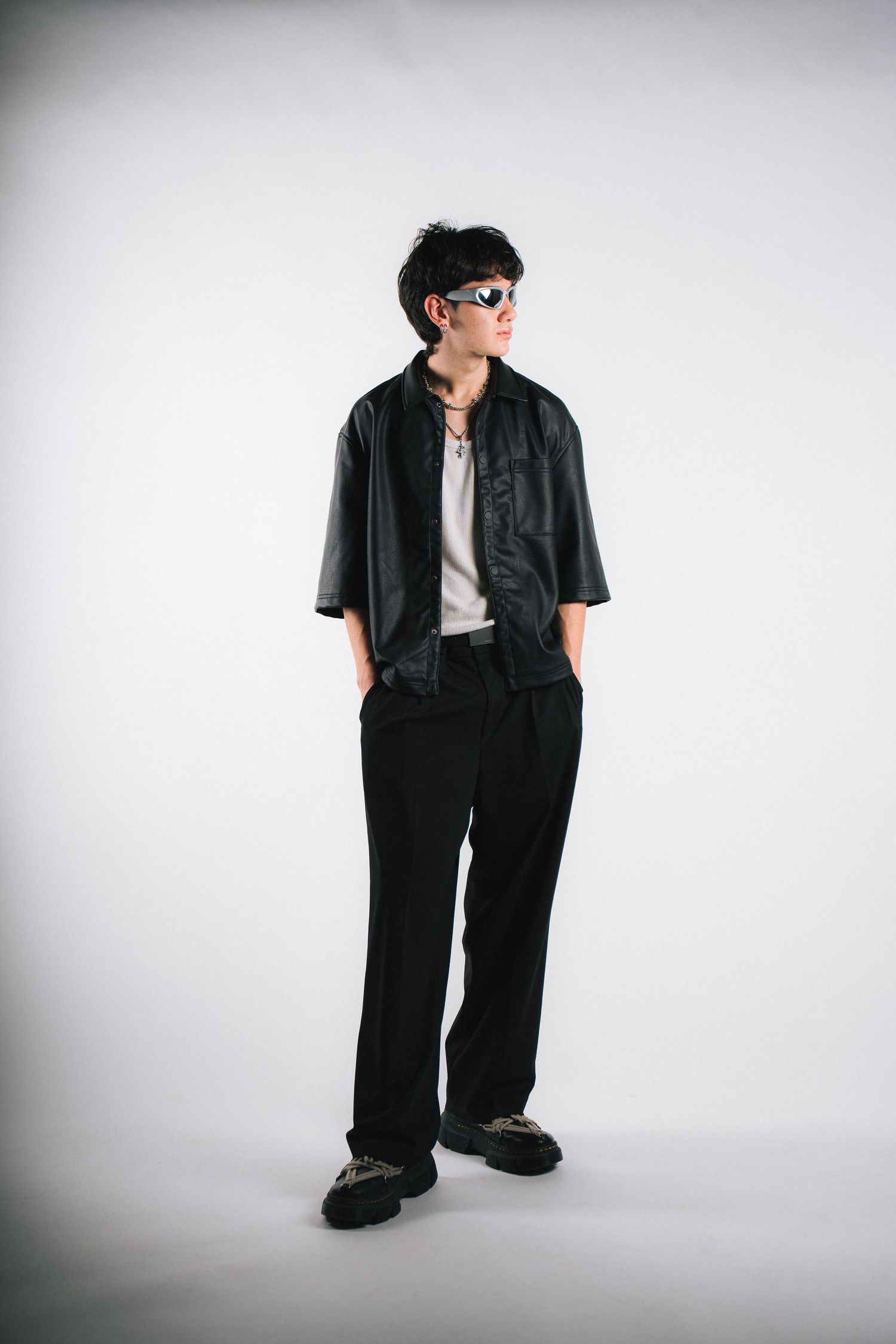 A person wearing a black short sleeve button-up shirt, white t-shirt, dark pants and sunglasses poses against a light backdrop.
