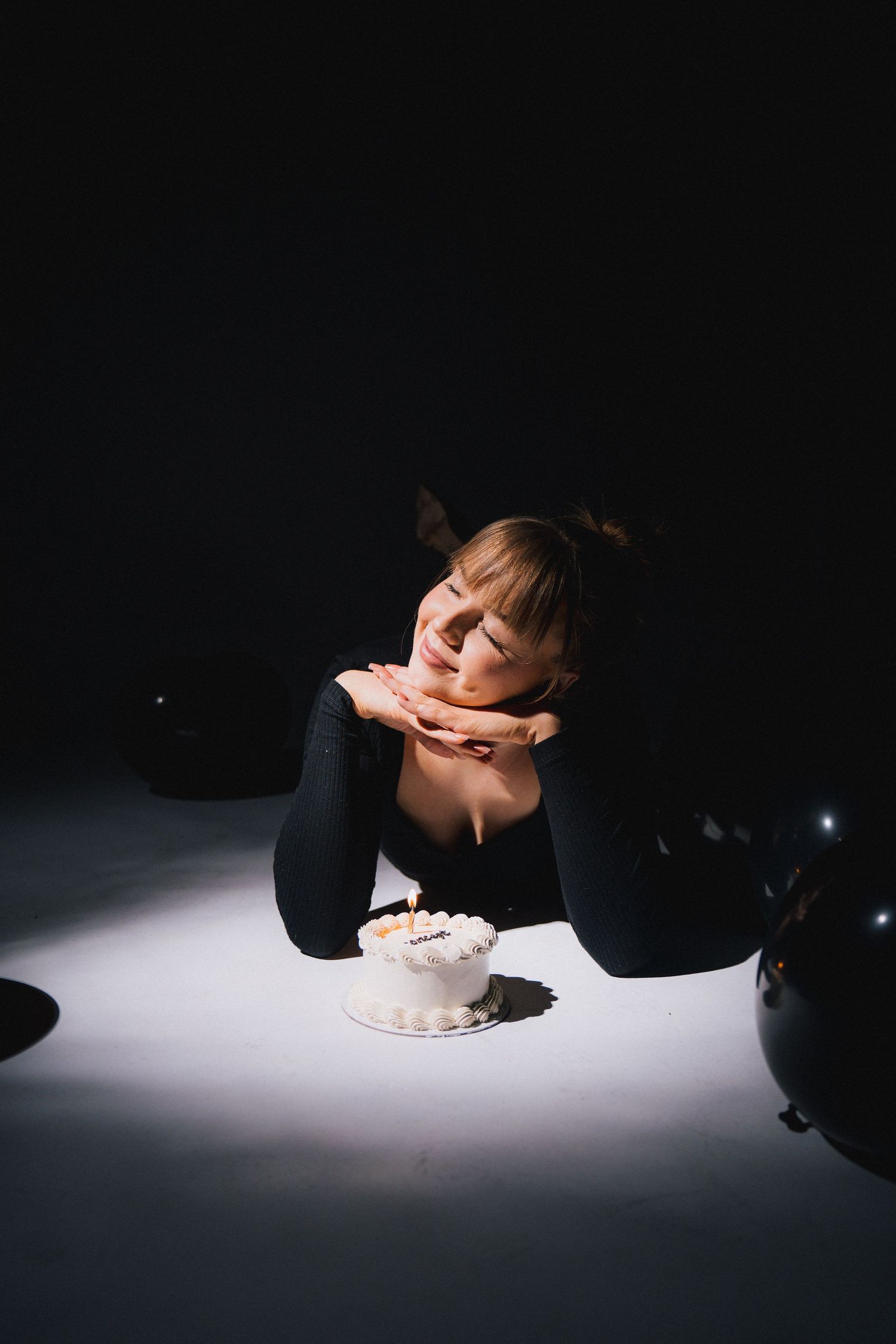 A person in black clothing lies on a white surface with a small white cake in moody dramatic lighting.
