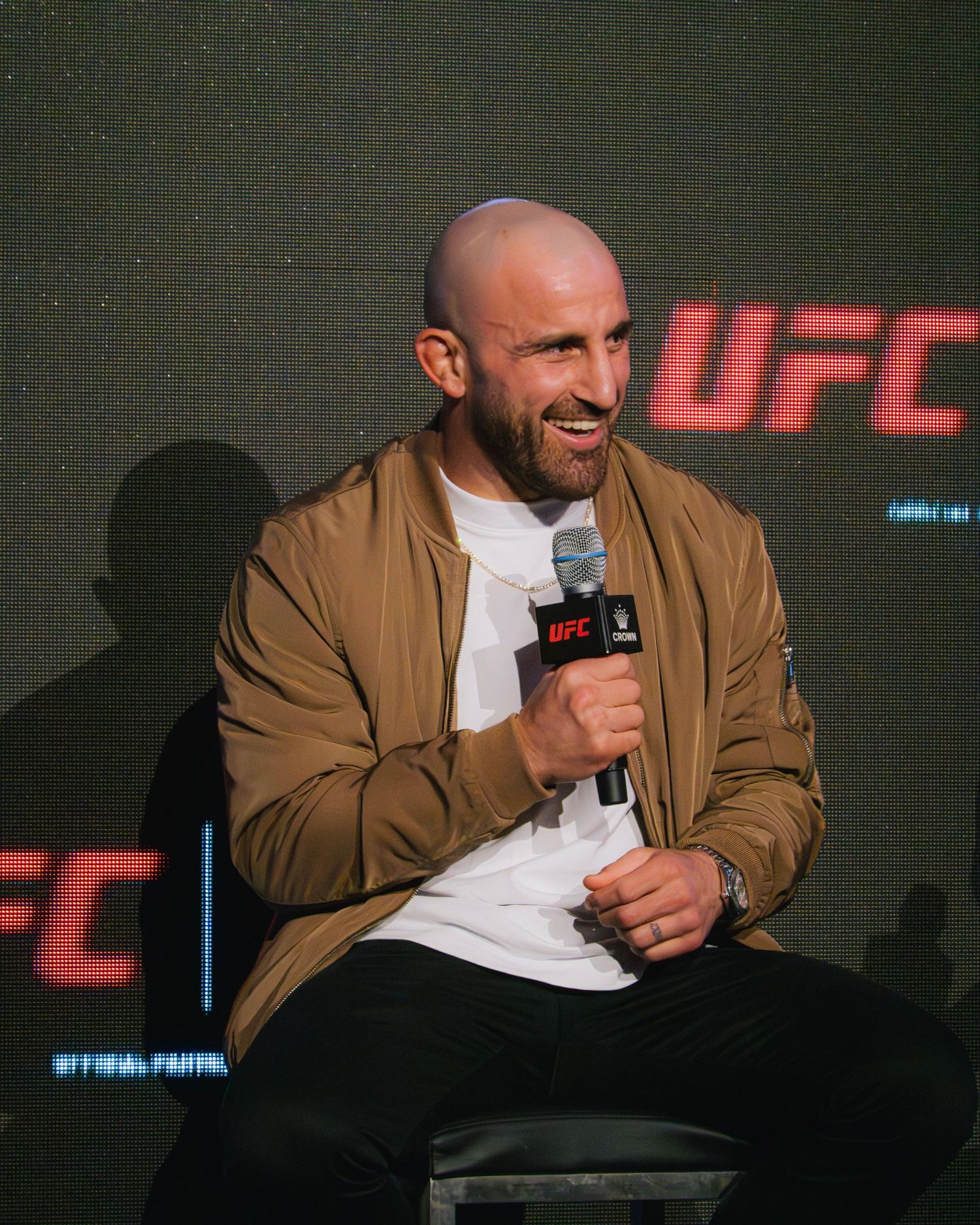 Fighter speaking into microphone at UFC press conference wearing brown jacket and white t-shirt.