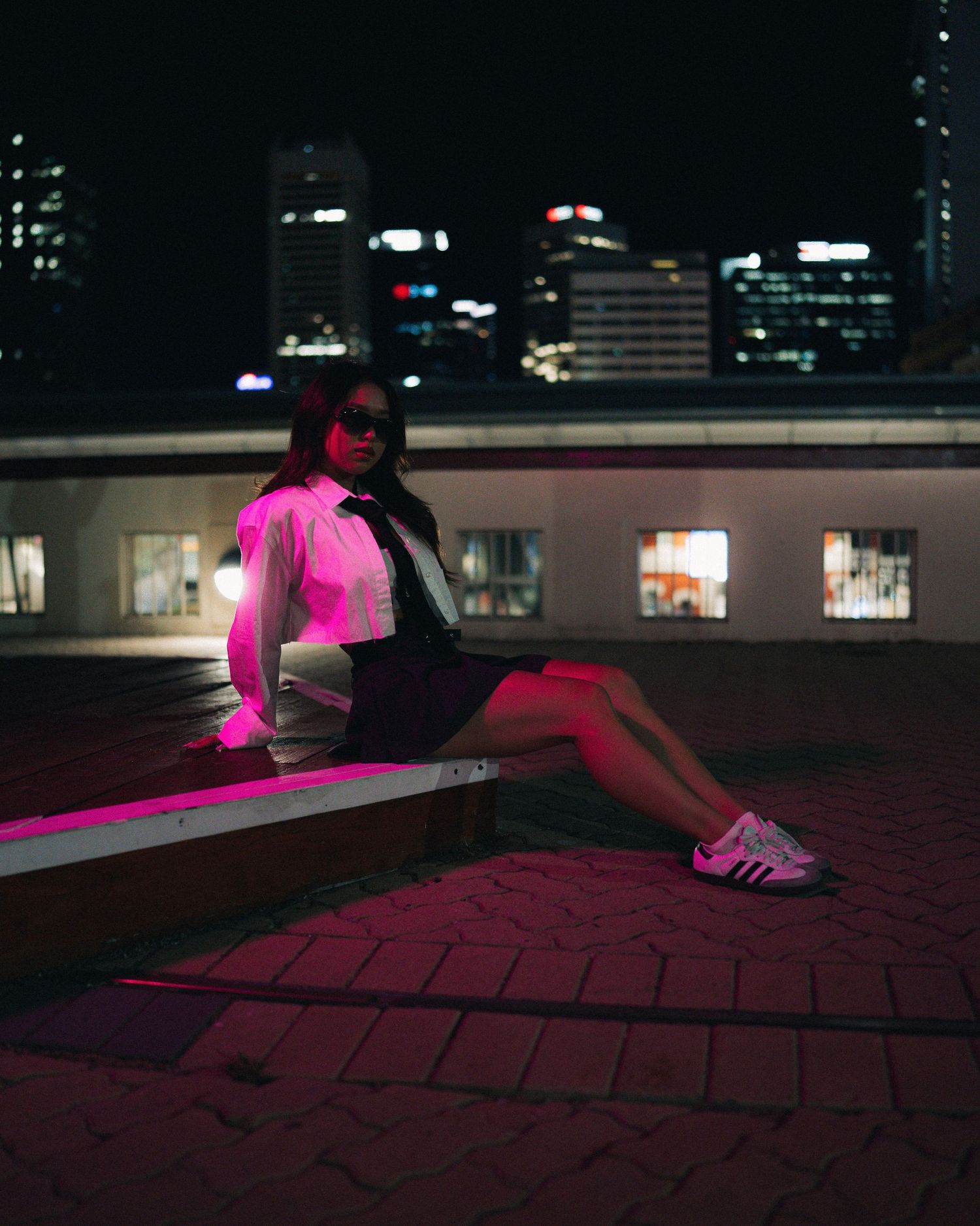 Person in pink illuminated clothing sits on urban rooftop at night with city lights behind.