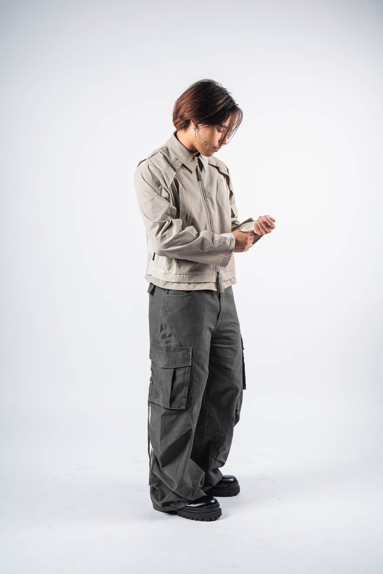 Person in beige jacket and gray cargo pants checking their watch against white background.