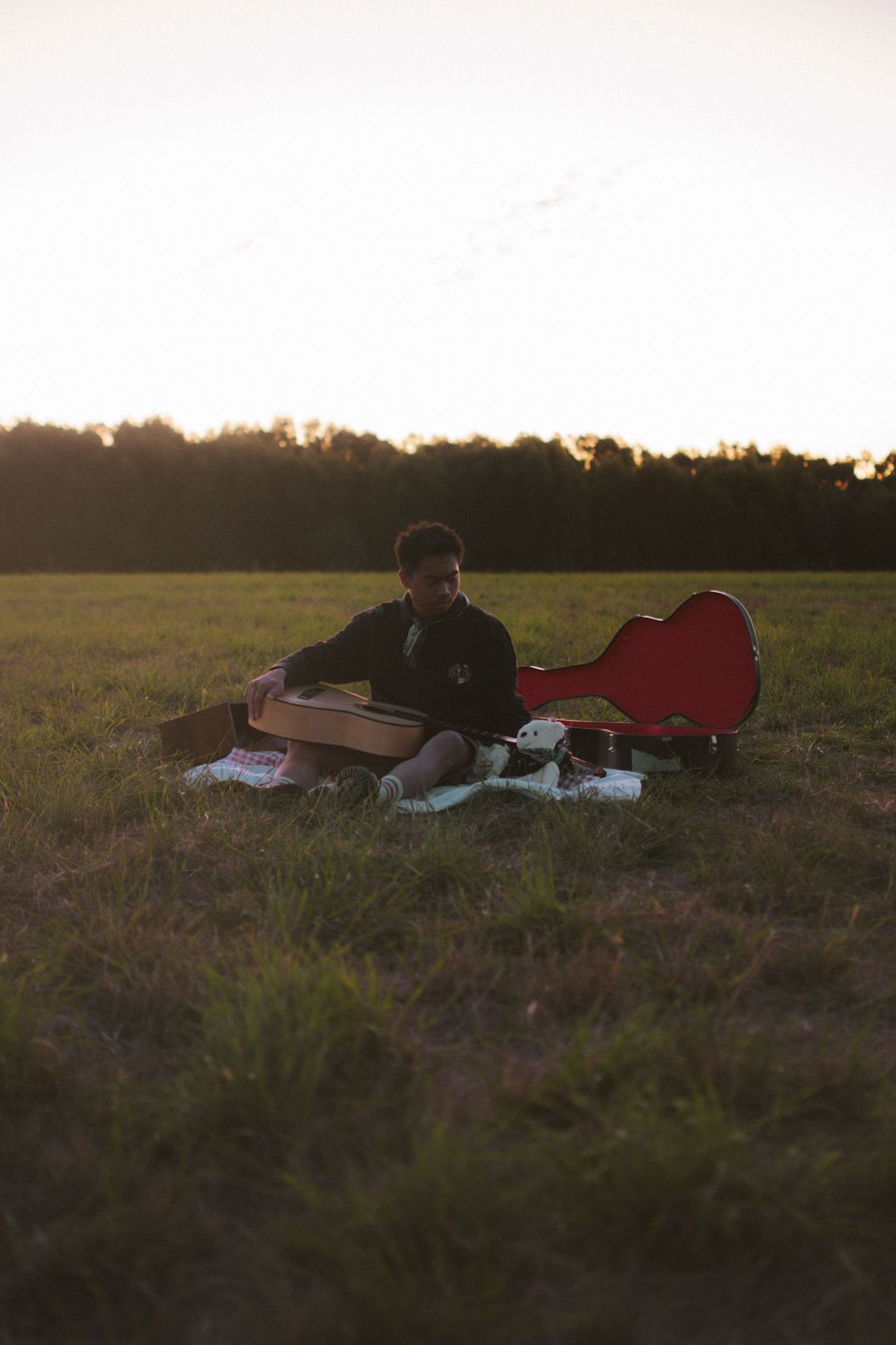 A person sits on a blanket in a field at sunset with a red guitar case and some papers spread around them.