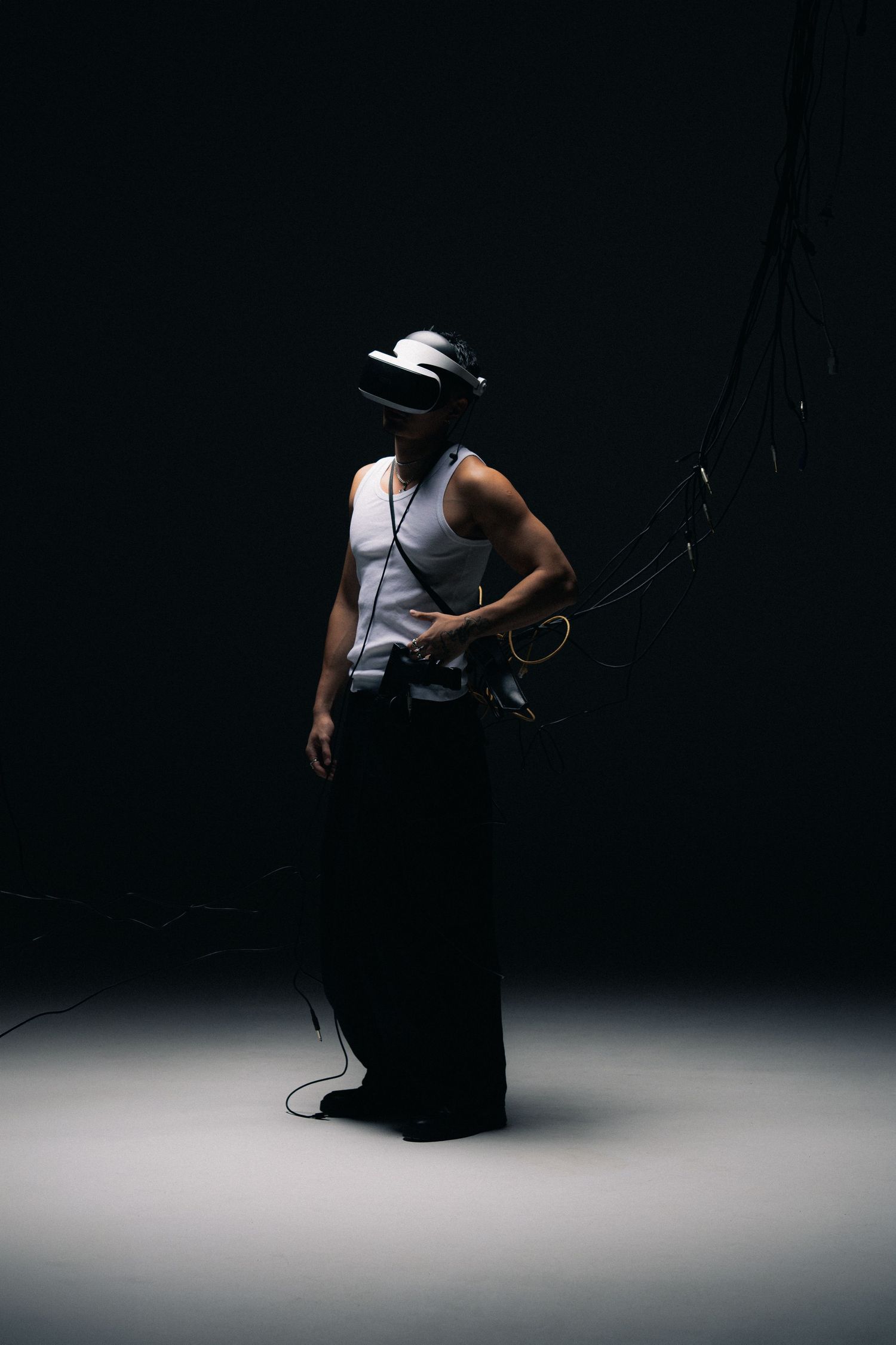 Figure in a white tank top and hat stands dramatically in a dark studio with dramatic spotlighting from above.