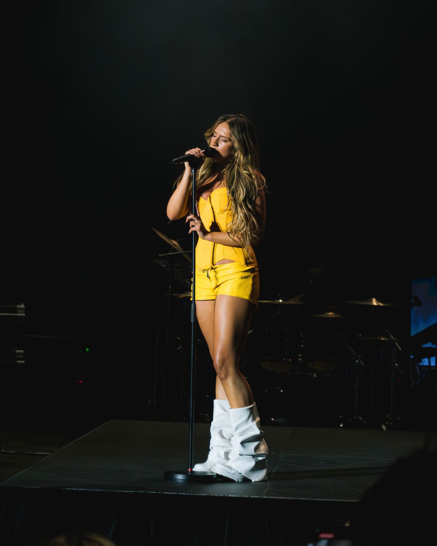 Performer in vibrant yellow dress and white boots sings on dark stage with microphone during concert performance.