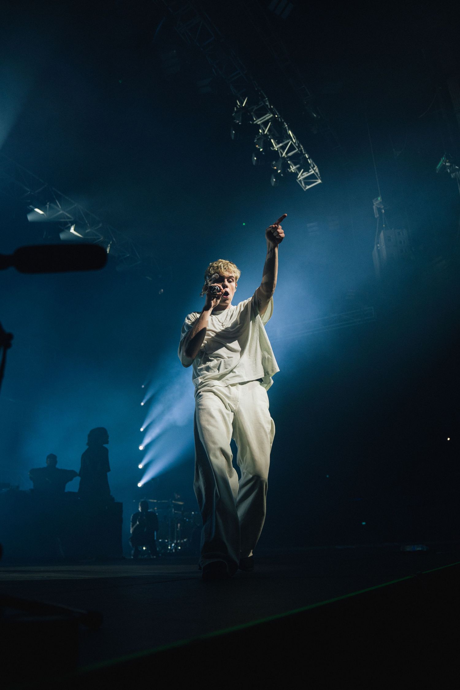 Performer in white outfit holds up microphone on atmospheric blue-lit stage during concert performance.