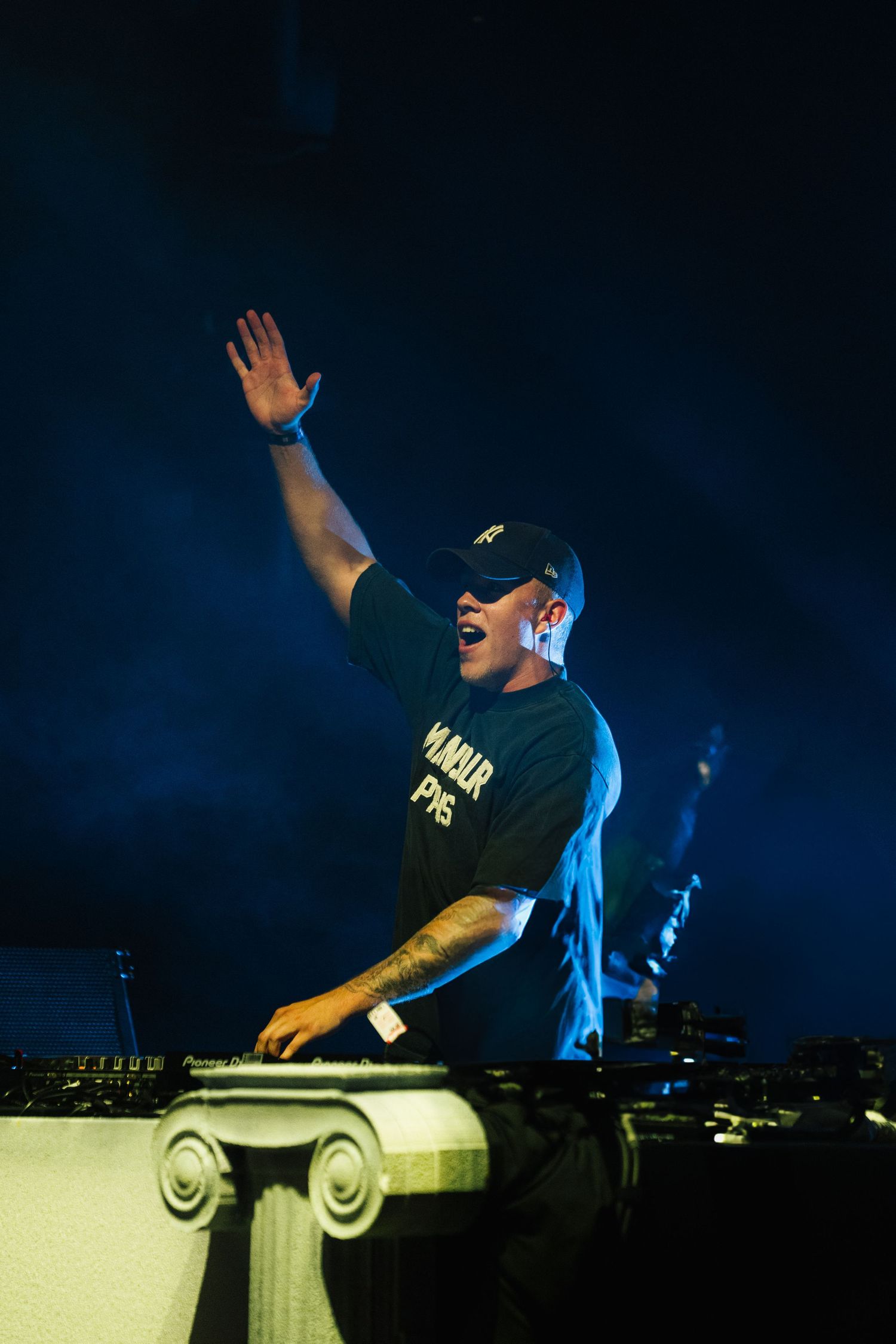DJ performing on elevated platform with raised hand under dramatic blue stage lighting.