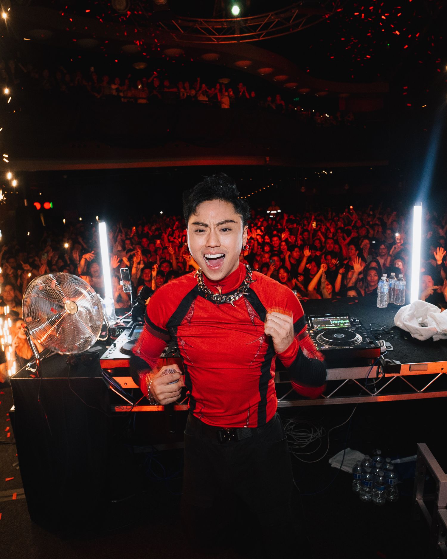 DJ in red outfit performs energetically behind decks with crowd visible in background.