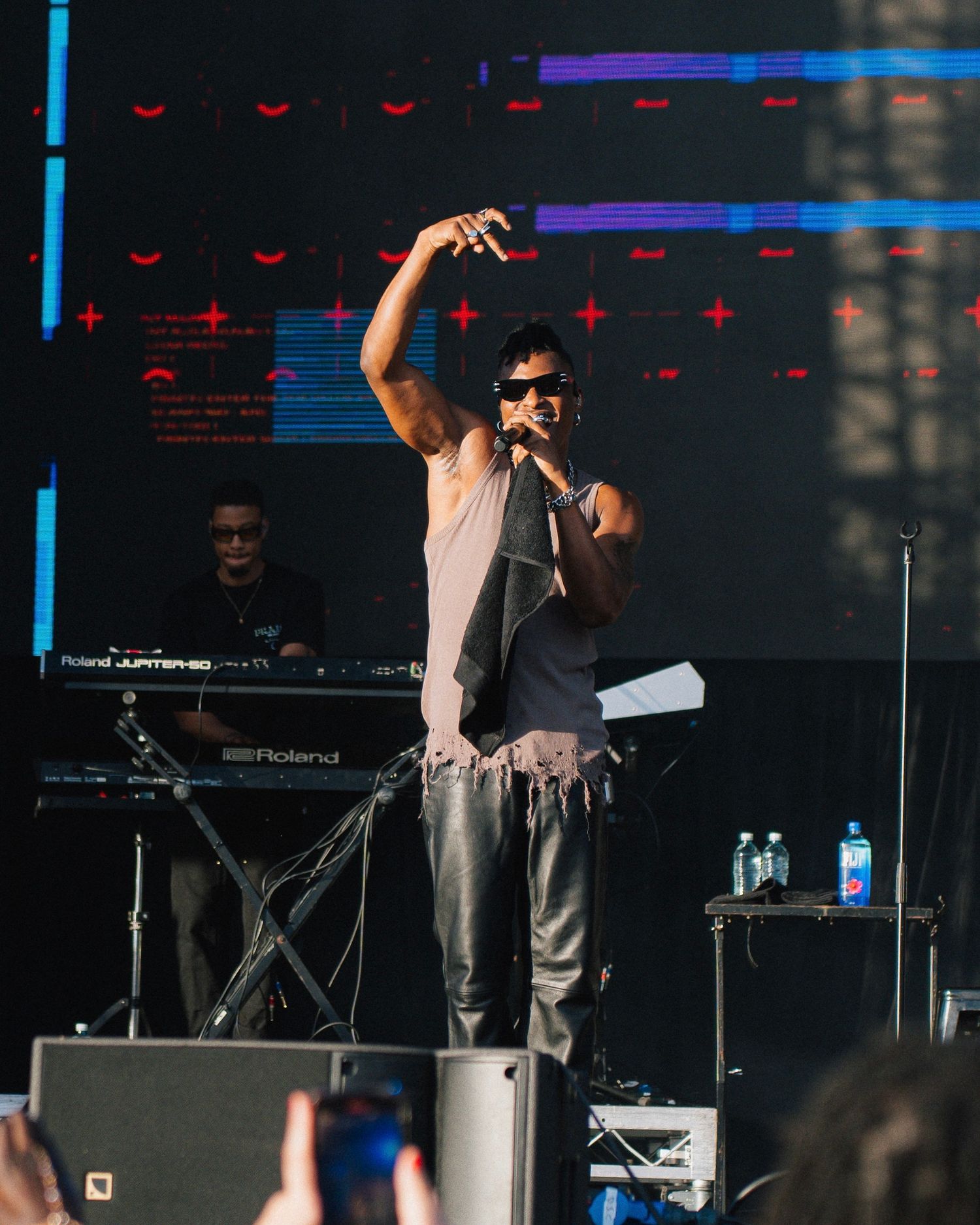 Concert performer in sleeveless shirt and sunglasses singing on stage with electronic equipment setup.