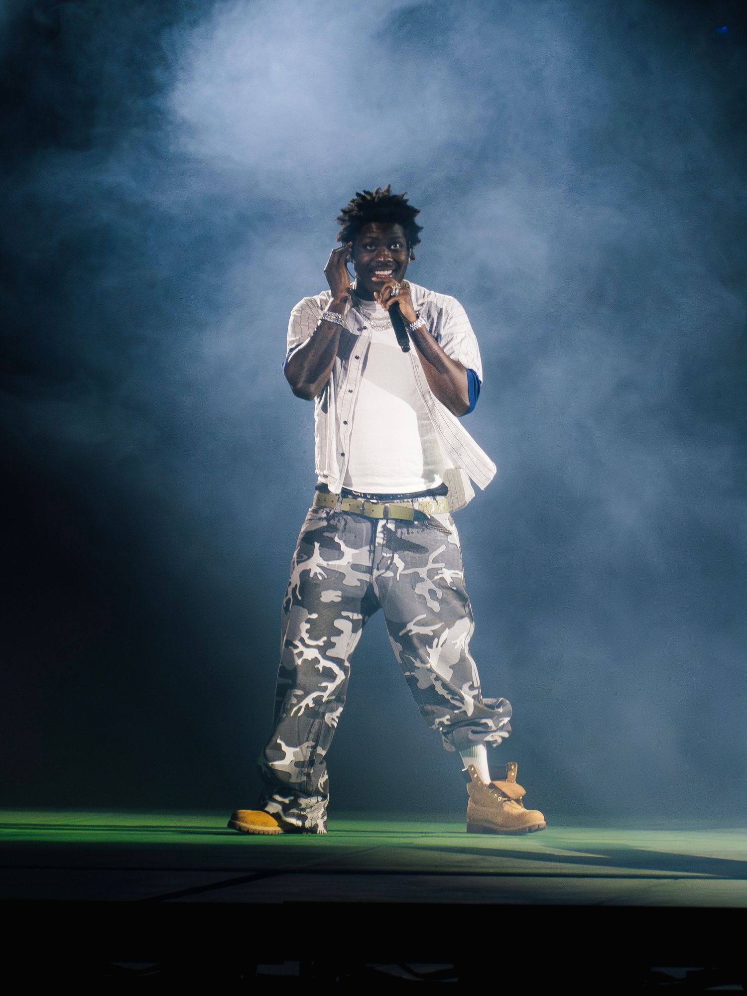 A performer in camo pants and white shirt stands on stage surrounded by atmospheric smoke effects and dramatic lighting.