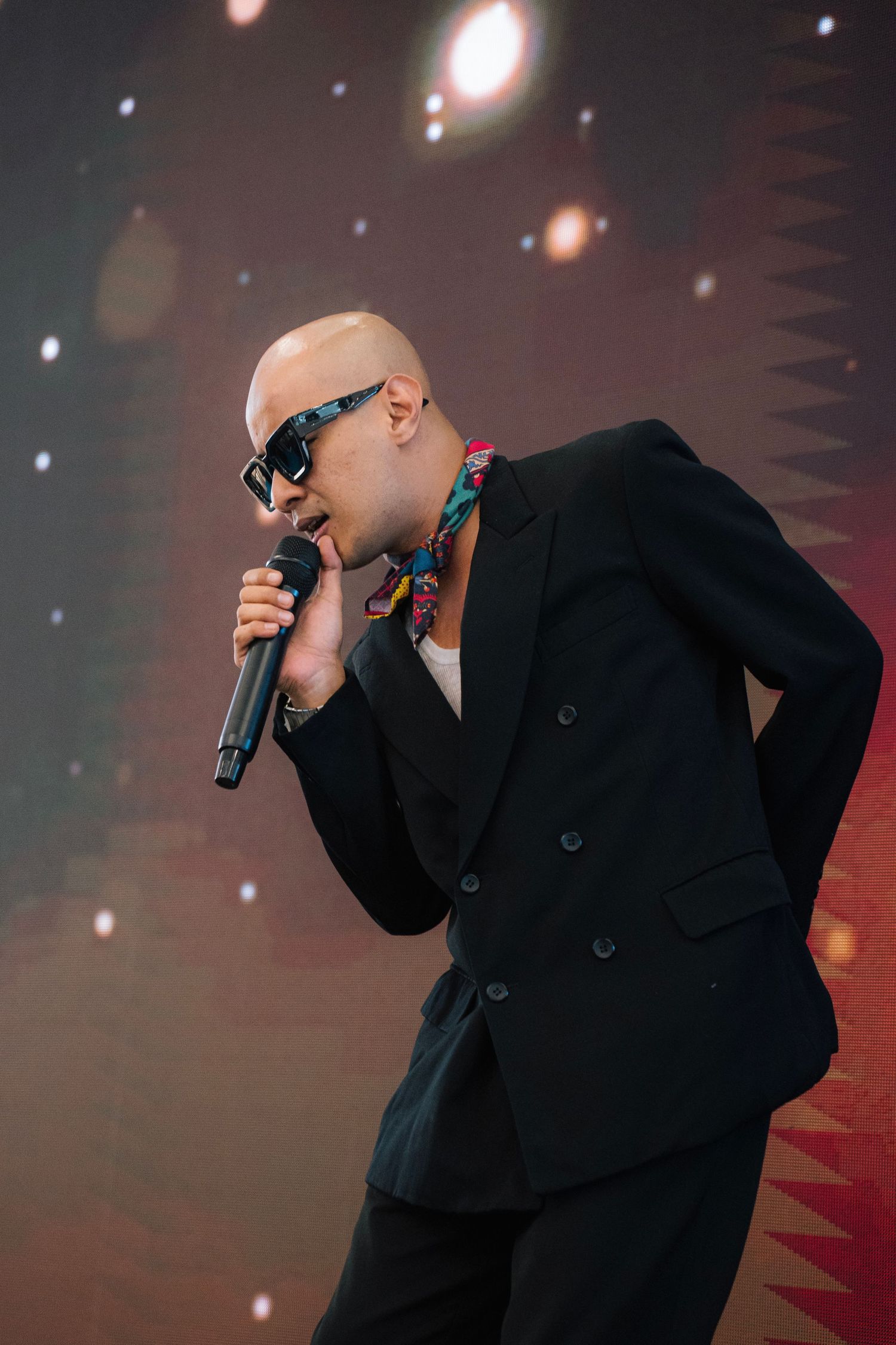 A performer in a black suit and sunglasses sings into a microphone on stage with bokeh lights in the background.