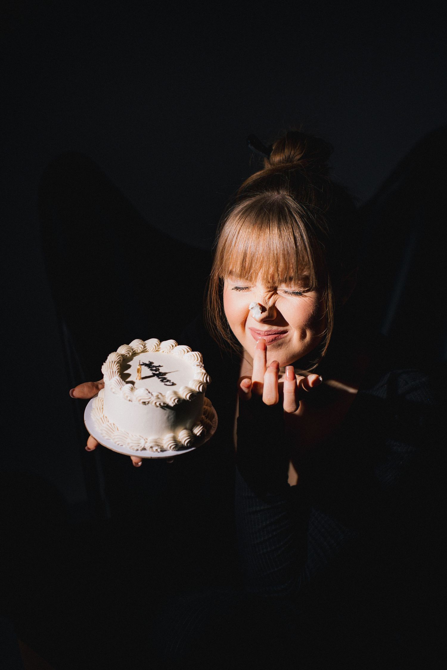 A whimsical portrait in dark lighting shows a person in black with a decorative white cake against a dark background.