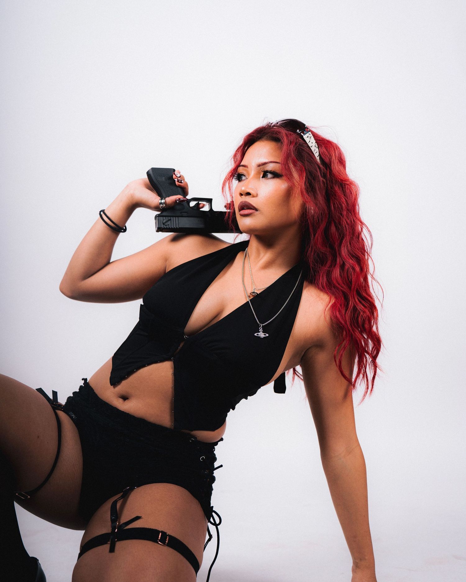 Model in edgy black outfit with bright red hair poses with gun prop against white background on fashion photoshoot.